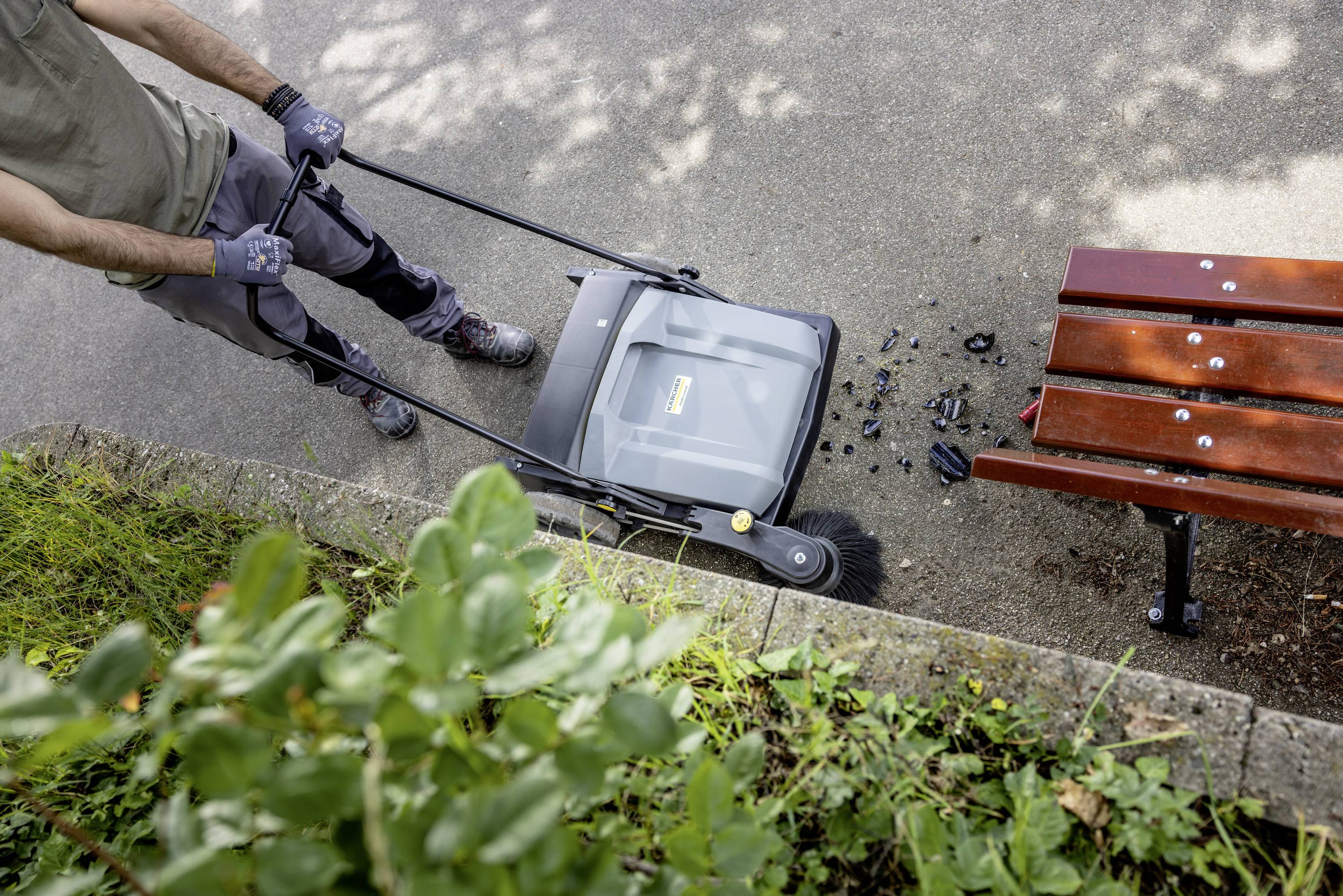 Osoba zamiata chodnik przy ławce za pomocą urządzenia do zamiatania. Na ziemi leżą liście i śmieci.