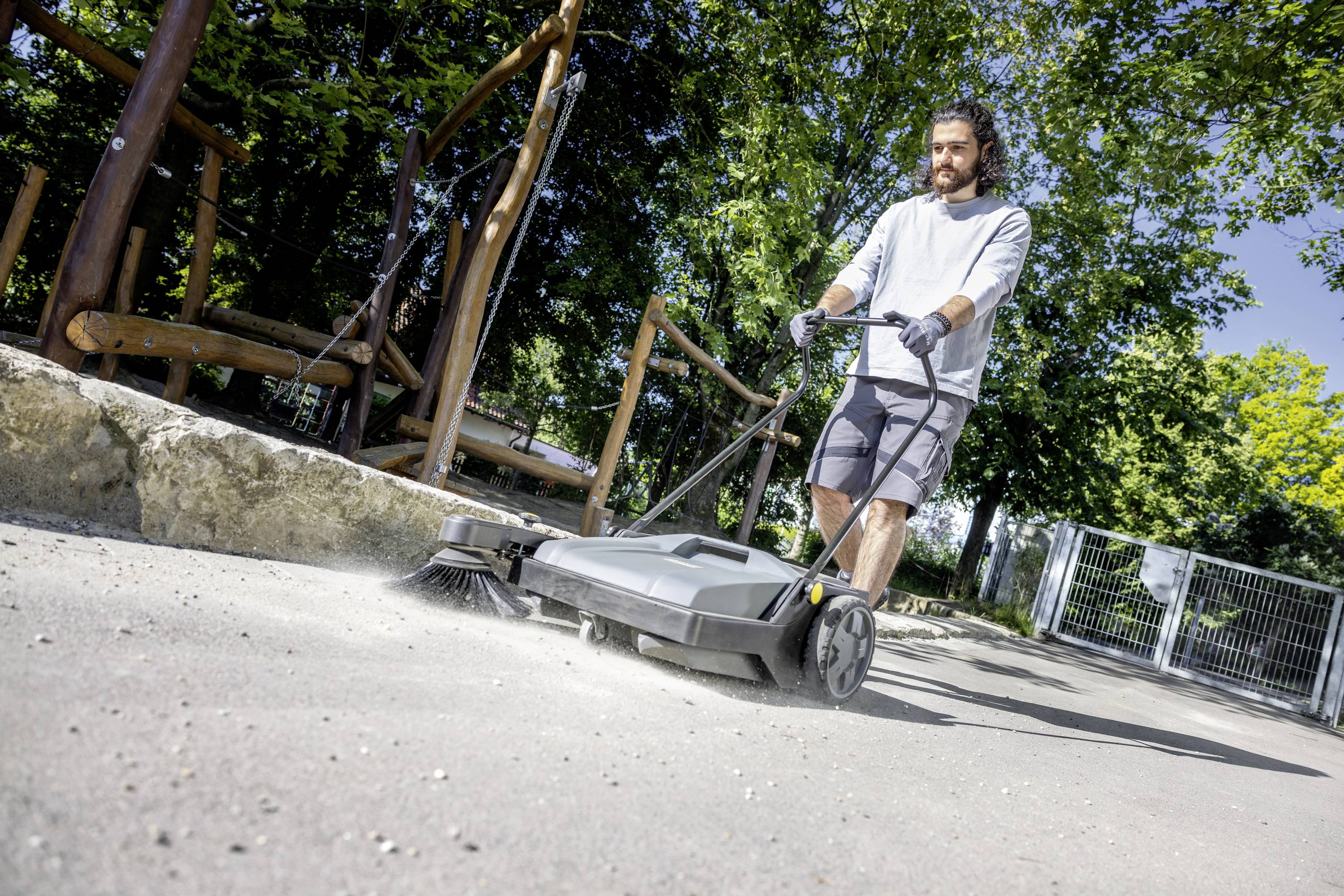 Mężczyzna zamiata chodnik za pomocą maszyny czyszczącej. W tle widać drzewa i ogrodzenie.