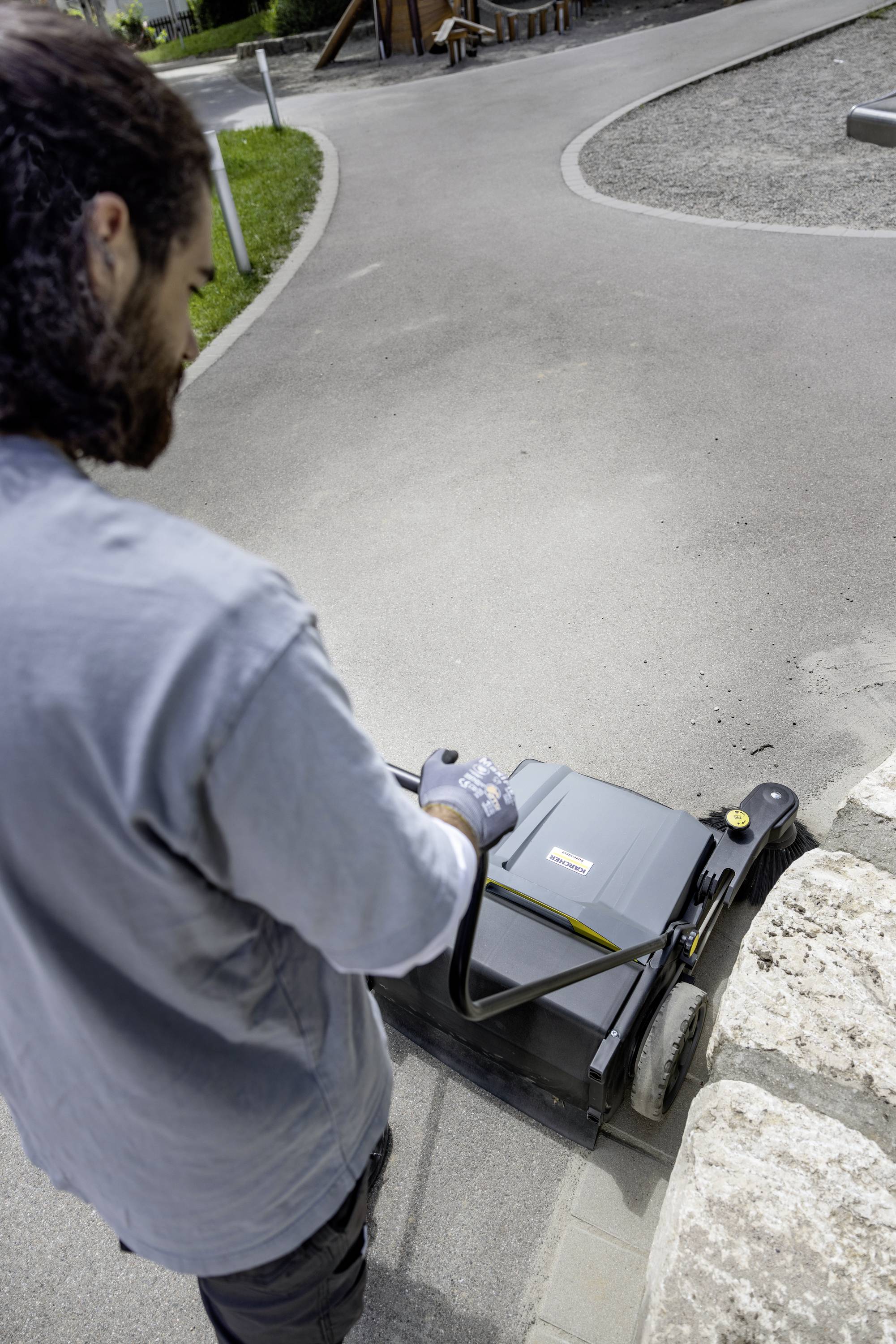 Osoba obsługuje zamiatarkę chodnikową przy murze. W tle widać obszar ogrodowy.