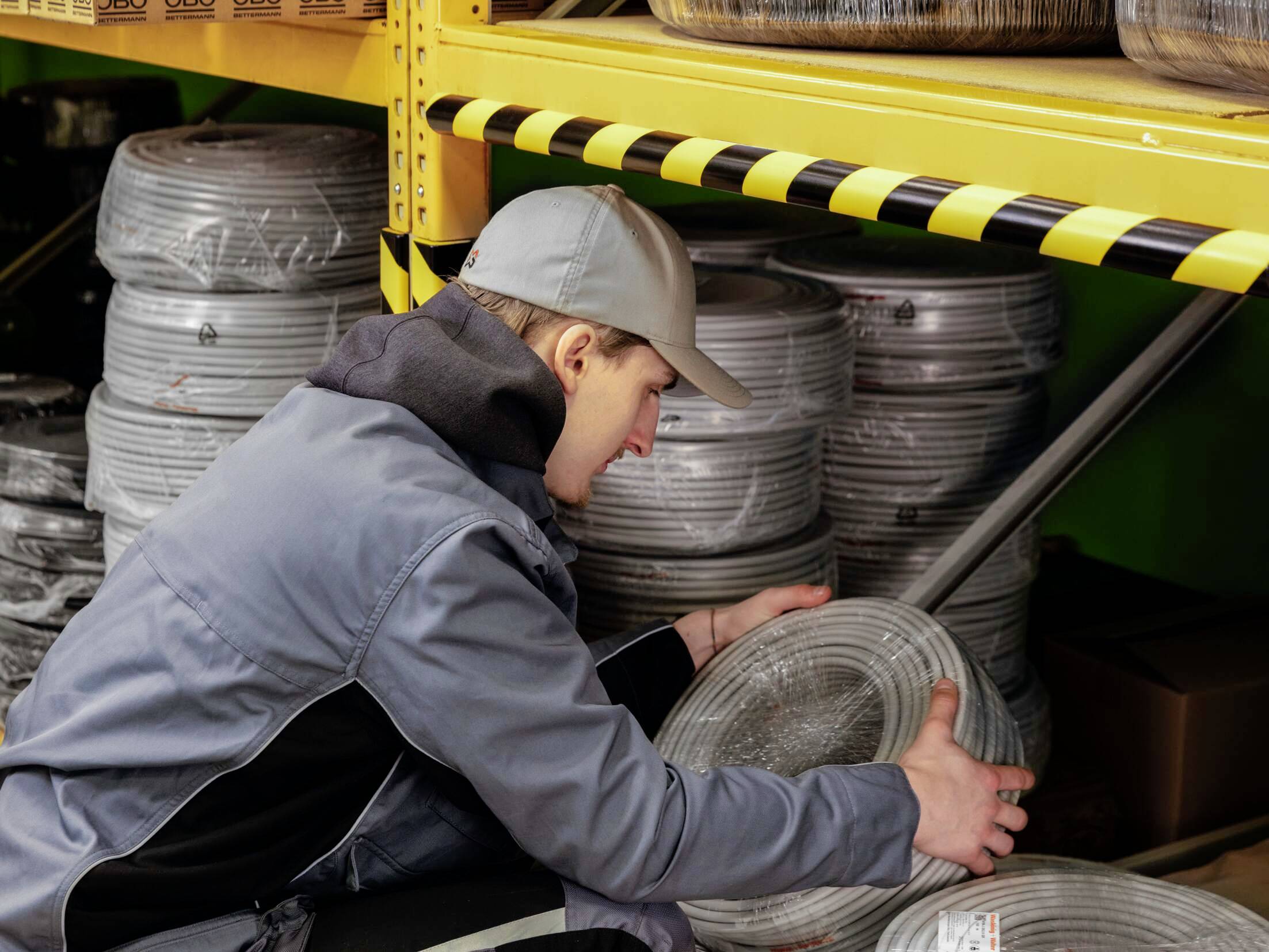 Mężczyzna w stroju roboczym porządkuje rolki kabli na żółtej półce magazynowej. Kable są zapakowane w plastik.