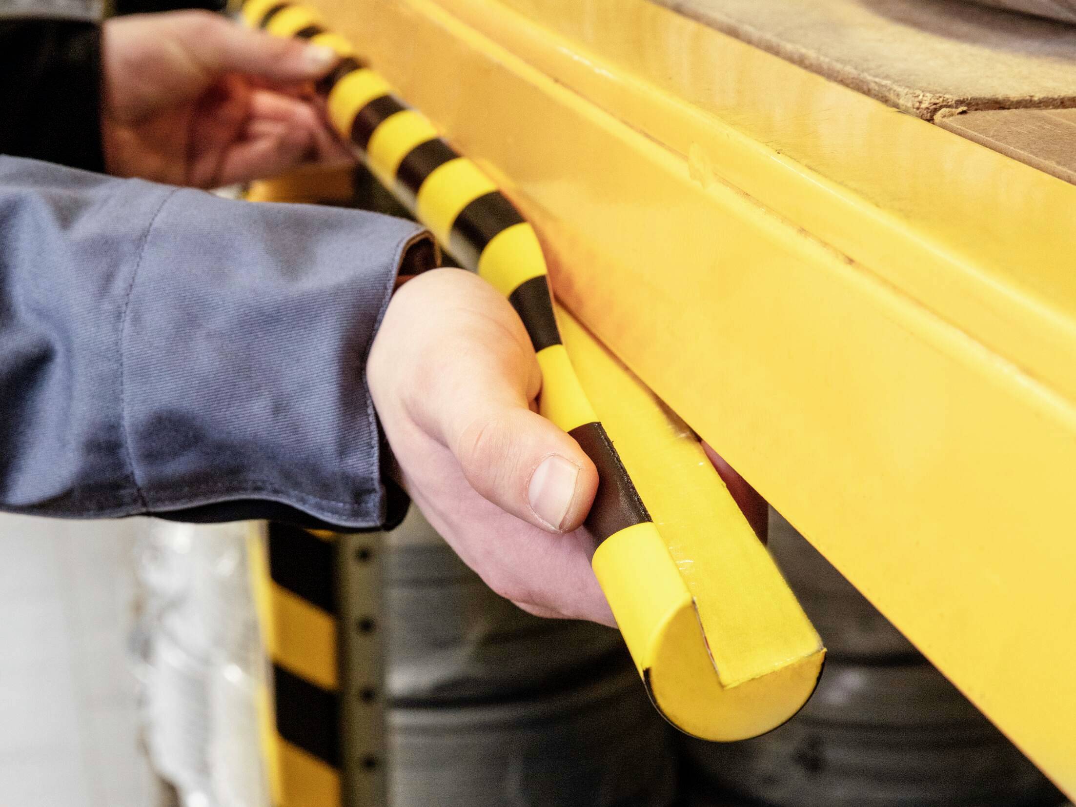 Osoba mocuje żółto-czarną taśmę ostrzegawczą na krawędzi regału w ramach środków bezpieczeństwa.