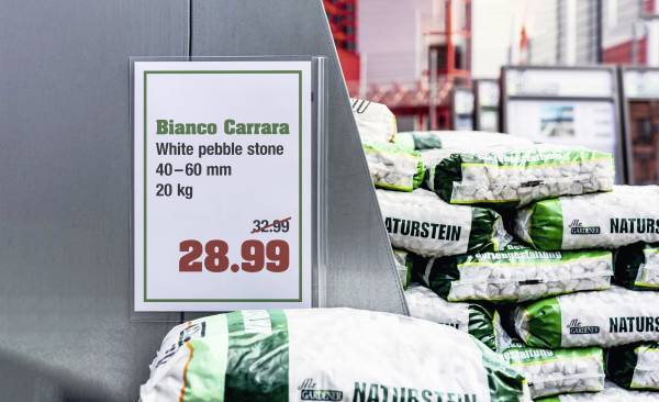 Kartka z ceną wskazuje 'Bianco Carrara', białe kamyki, 40-60 mm, 20 kg, za 28,99 EUR. W tle widać ułożone worki.