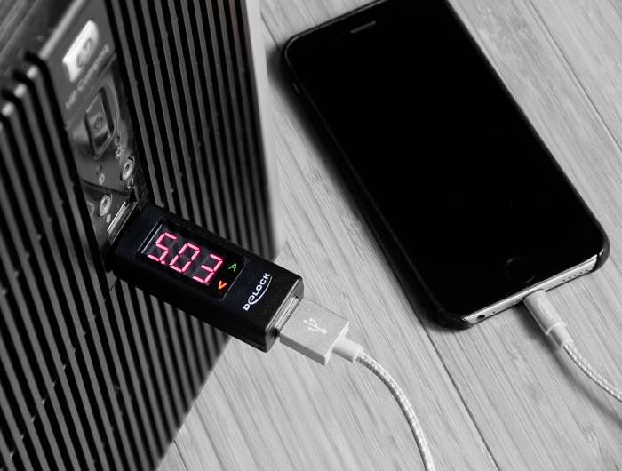 Czarny smartfon jest podłączony do ładowarki za pomocą kabla USB. Miernik wskazuje napięcie '5.0 V'.