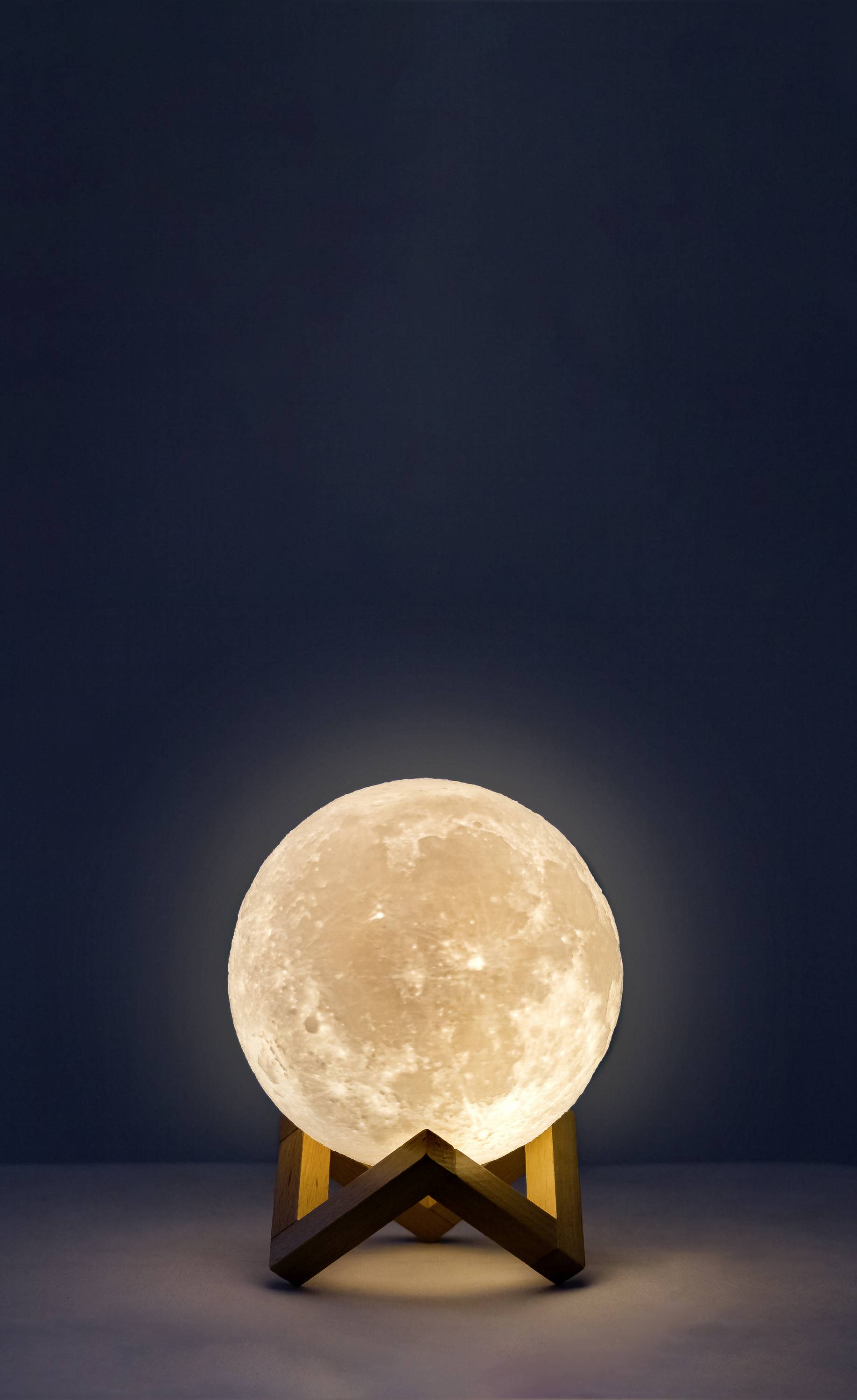 Oświetlona lampa w kształcie księżyca stoi na drewnianym stojaku, otoczona ciemnością, symbolizując nocny spokój i kosmiczną estetykę.