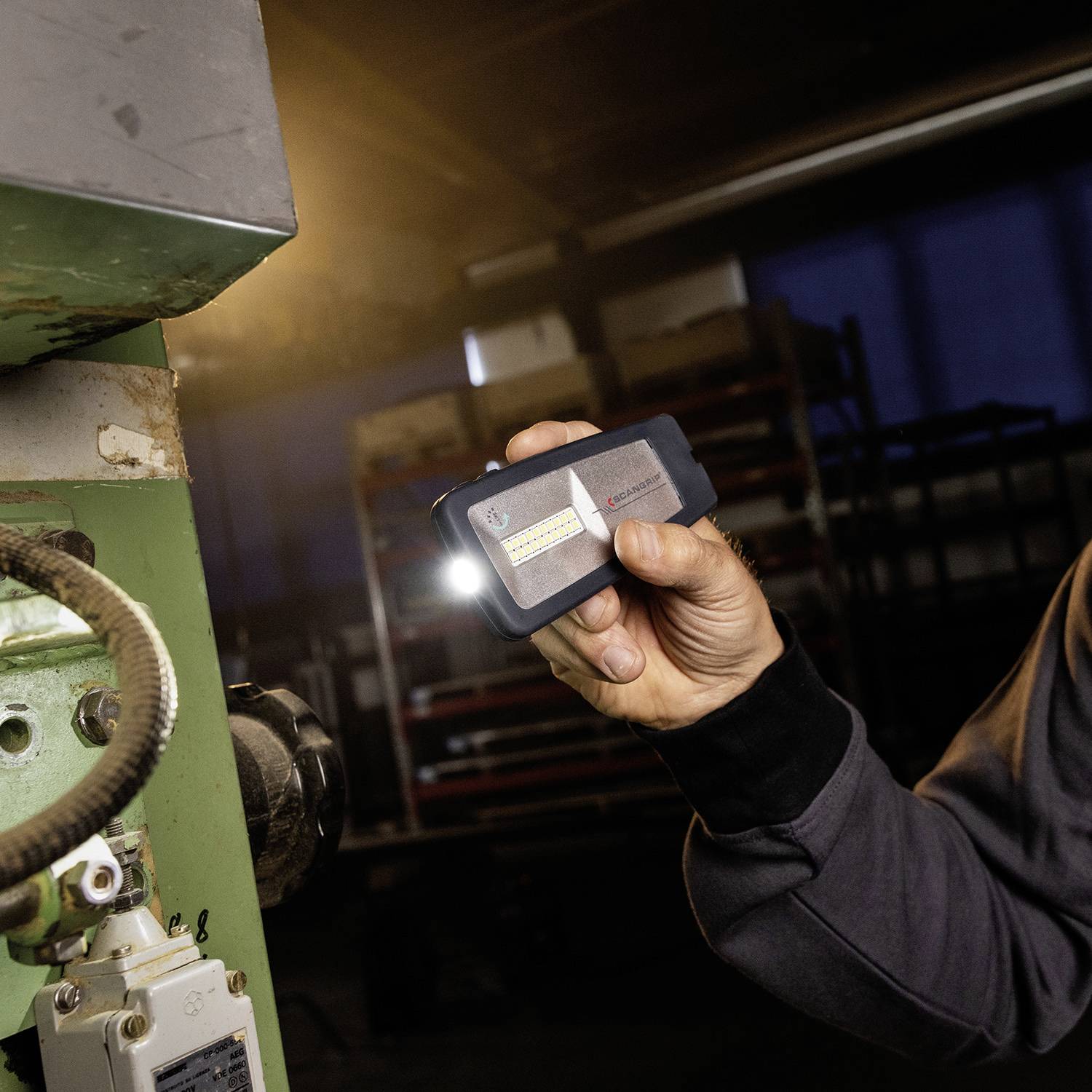 Osoba trzyma w ręku małe, prostokątne urządzenie pomiarowe ze świecącą lampką. Otoczenie sprawia wrażenie przemysłowego.