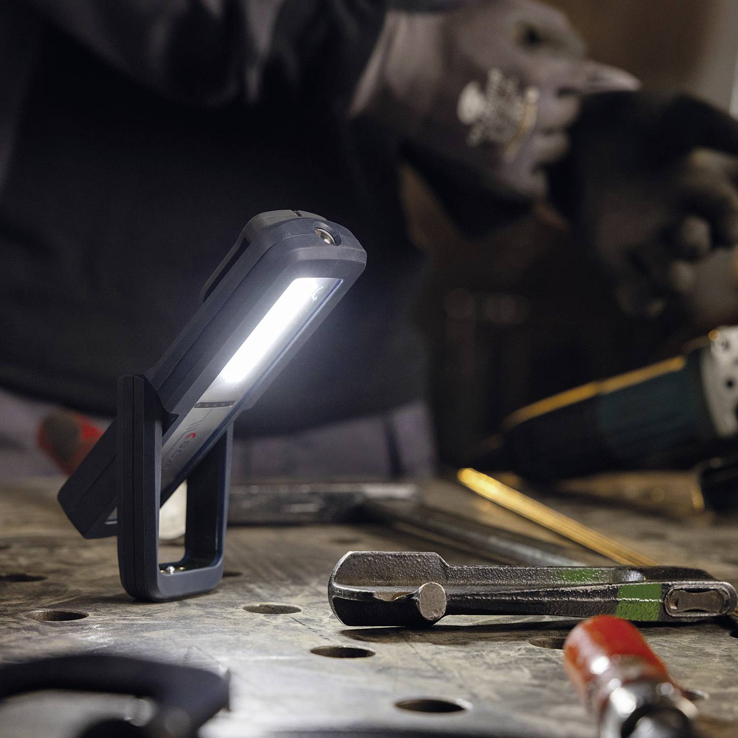 Robotnik obsługuje narzędzia na stole warsztatowym, podczas gdy przenośna lampa robocza oświetla powierzchnię.
