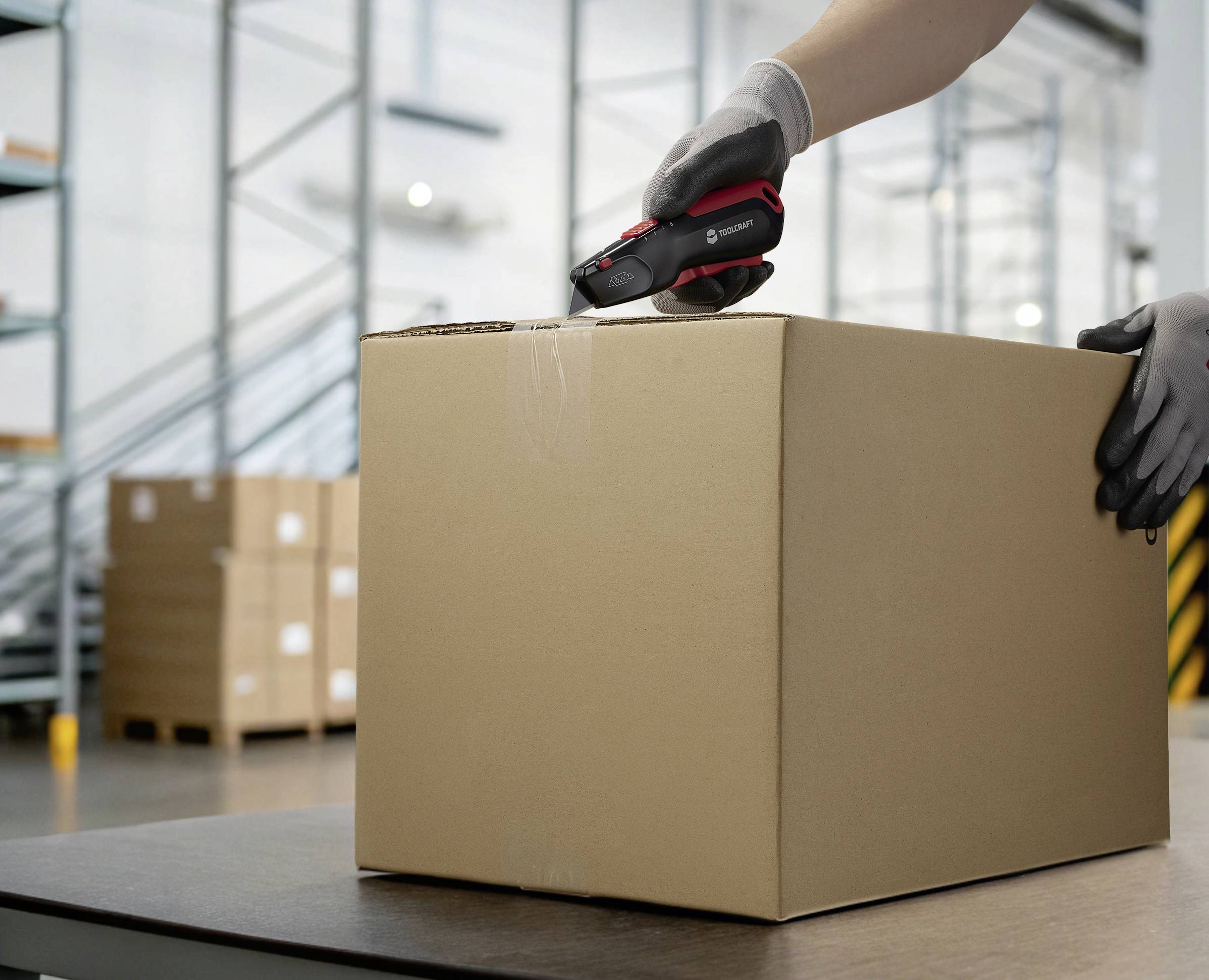 Osoba otwiera duży brązowy karton czarno-czerwonym nożem monterskim w magazynie z regałami.
