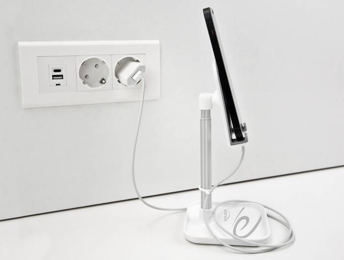 Tablet stoi na stojaku i jest podłączony kablem ładującym do gniazdka elektrycznego. Po lewej stronie widoczne są dwa gniazdka elektryczne oraz dwa porty USB.