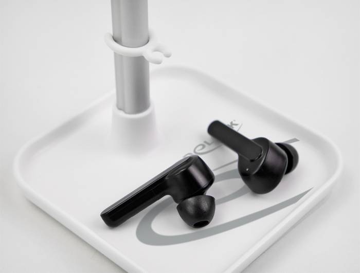 Czarne bezprzewodowe słuchawki douszne leżą na białym uchwycie. Metalowa rurka wznosi się pionowo z podstawy.