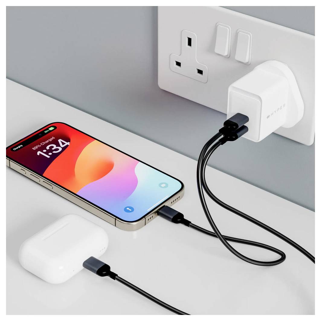 Smartfon ładuje się, podłączony do ładowarki w gniazdku ściennym. Obok leży para bezprzewodowych słuchawek.