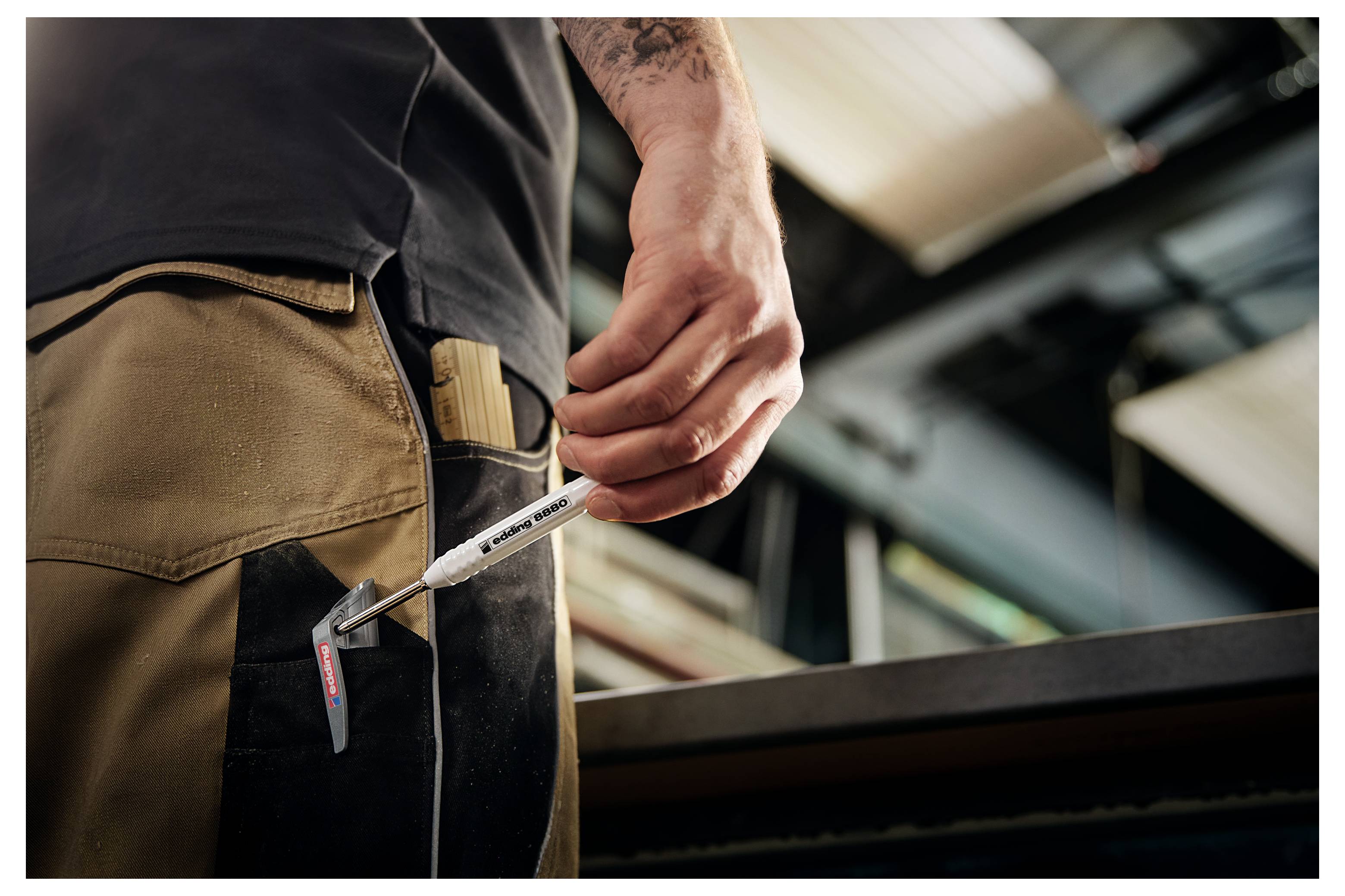 Zbliżenie na rękę pracownika trzymającego metalowe narzędzie przytwierdzone do paska obok beżowych spodni roboczych w ustawieniu przemysłowym.