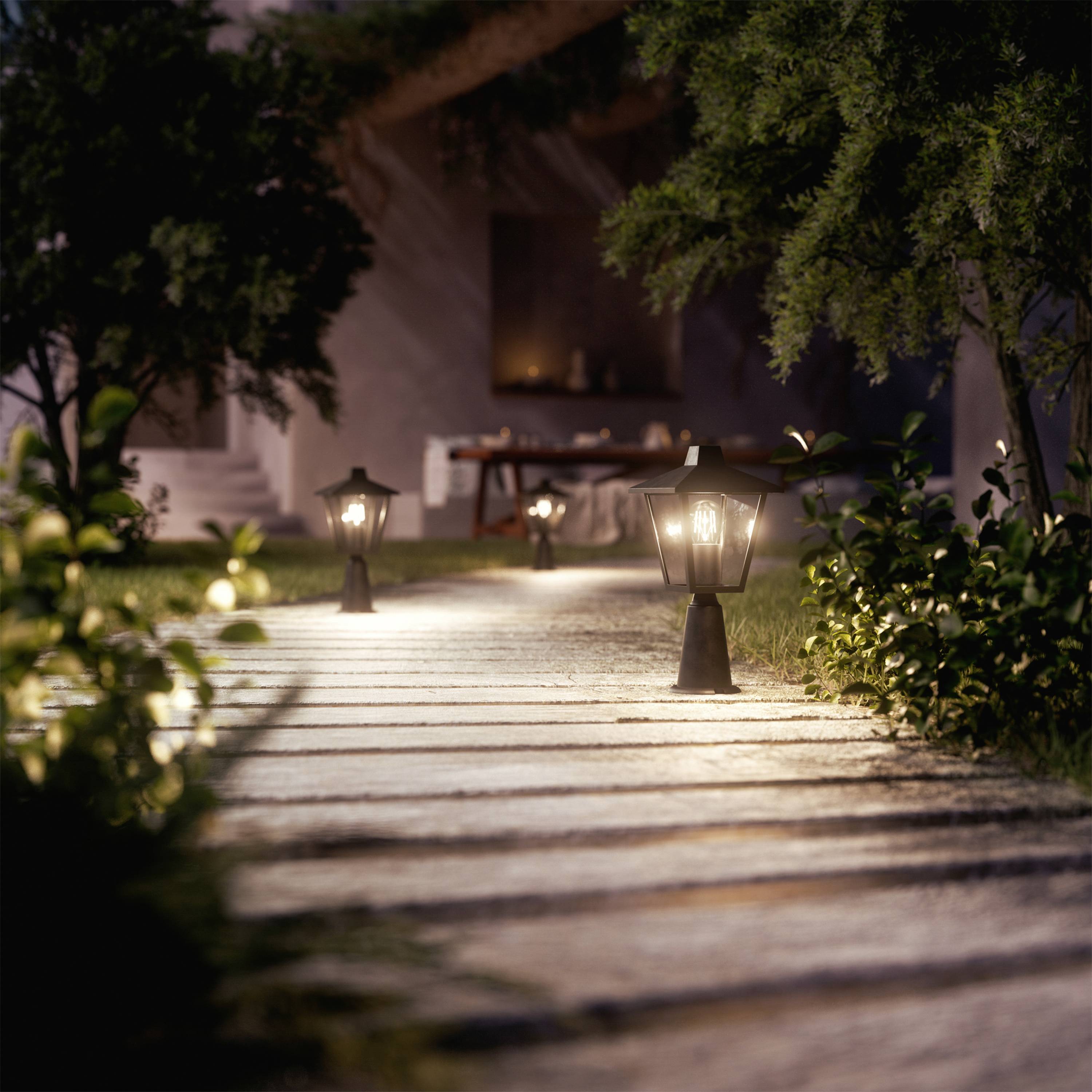 Ścieżka ogrodowa z latarniami, które wieczorem się świecą. W tle siedzi osoba przy drewnianym stole przed domem.