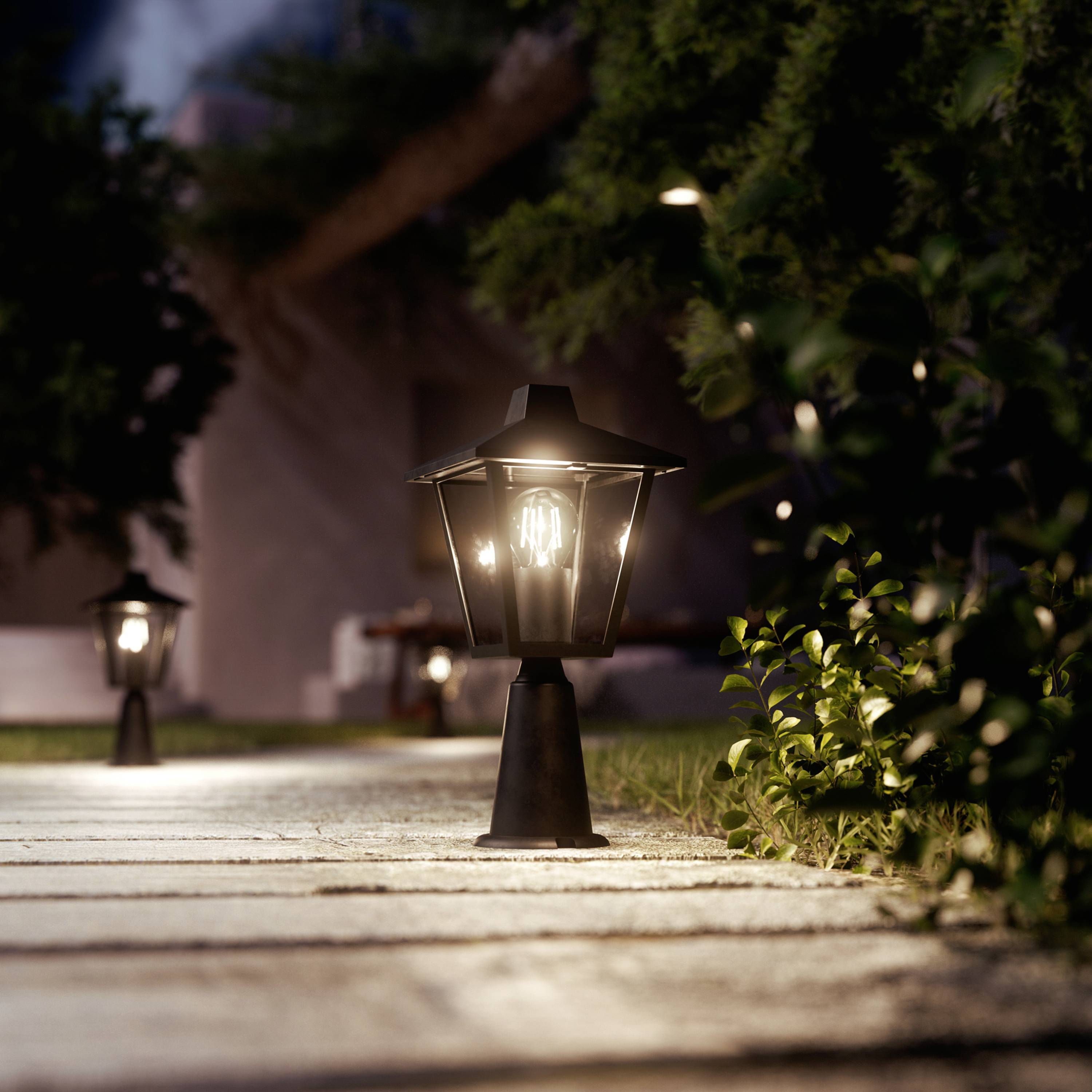 Wybrukowana ścieżka ogrodowa jest oświetlona kilkoma dekoracyjnymi latarniami o zmierzchu, otoczona drzewami i krzewami.