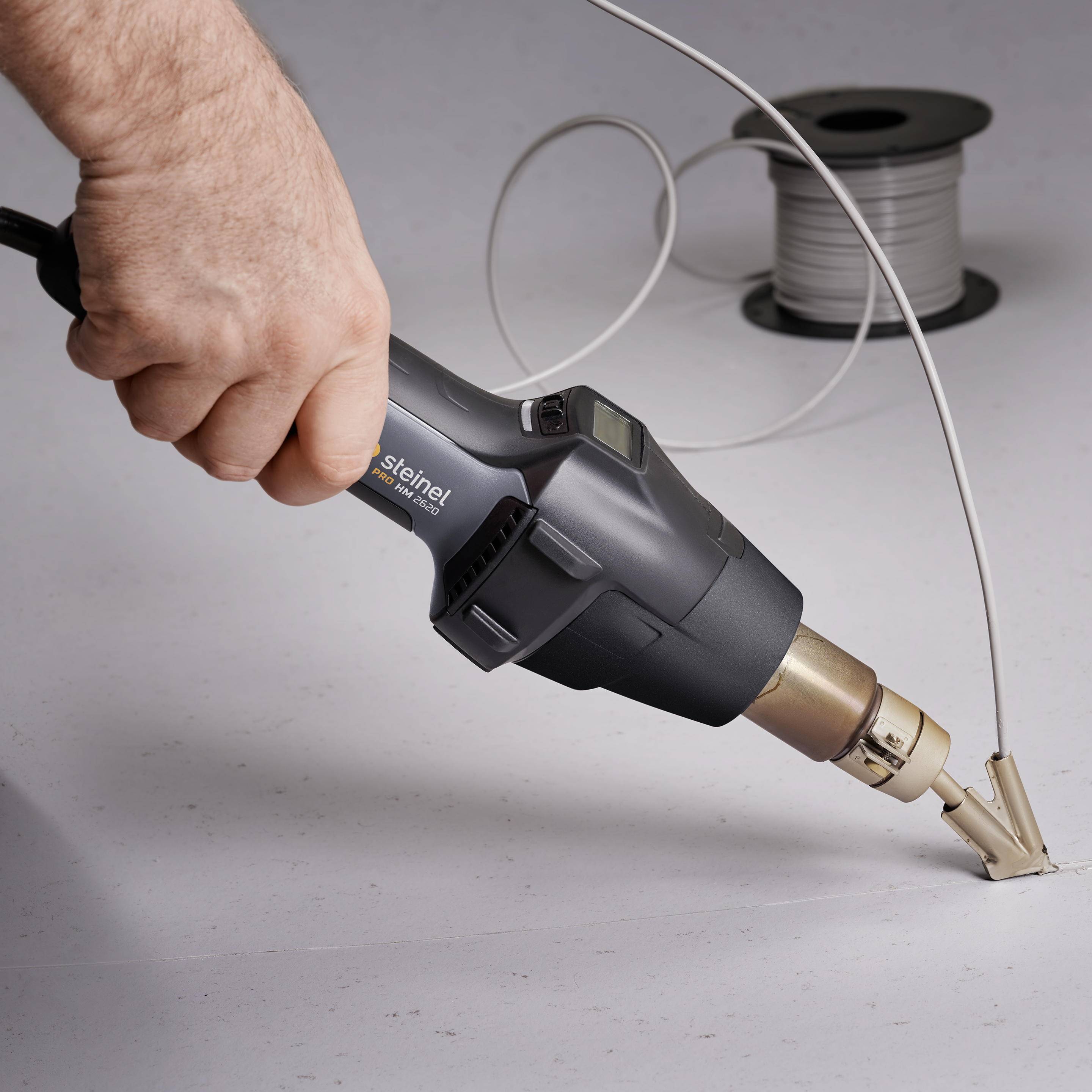 Jedna ręka trzyma pistolet na gorące powietrze skierowany na fragment przewodu, aby go termicznie obkurczyć. W tle rolka kabla.