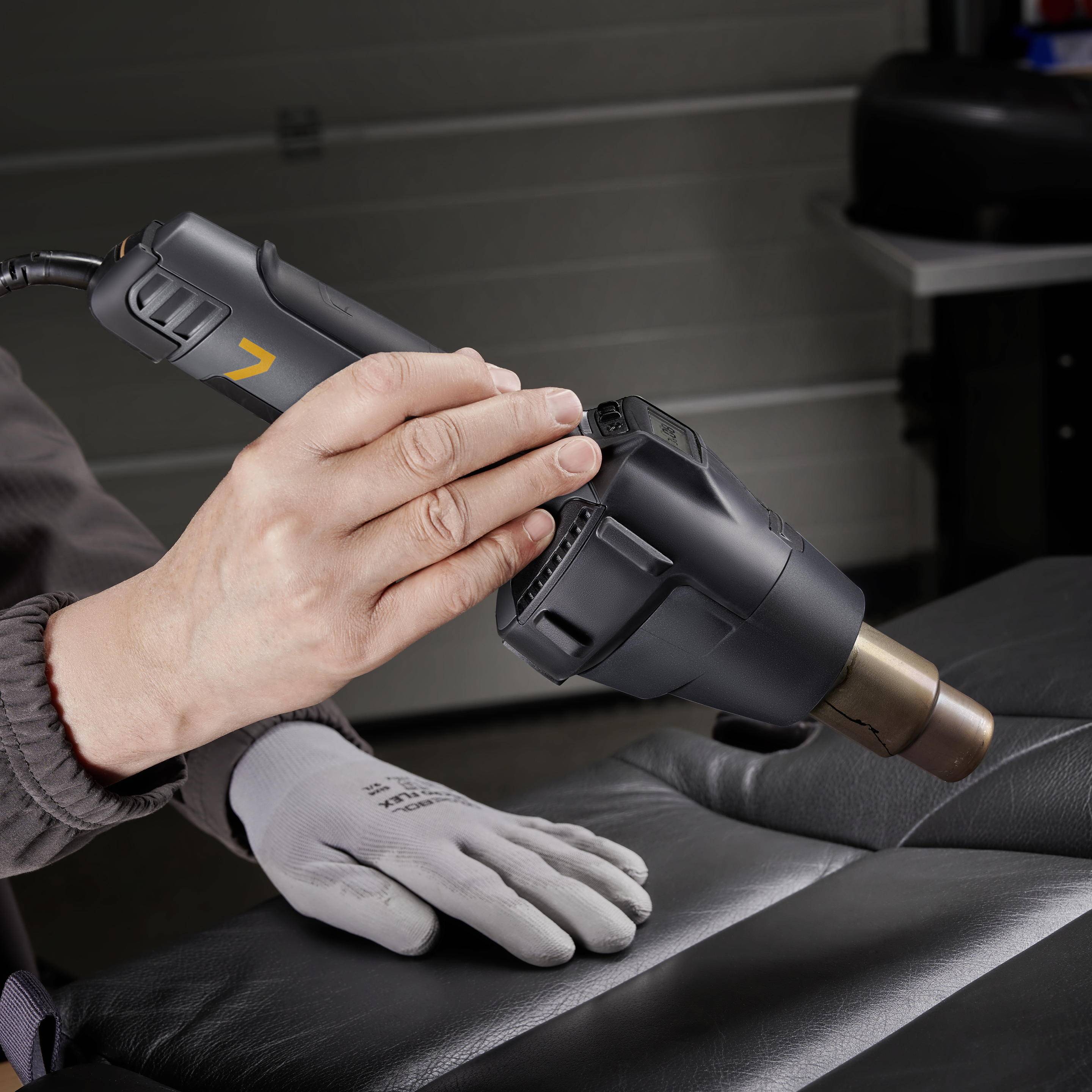 Osoba trzyma czarny pistolet gorącego powietrza, prawdopodobnie podczas pracy przy tapicerce w warsztacie.