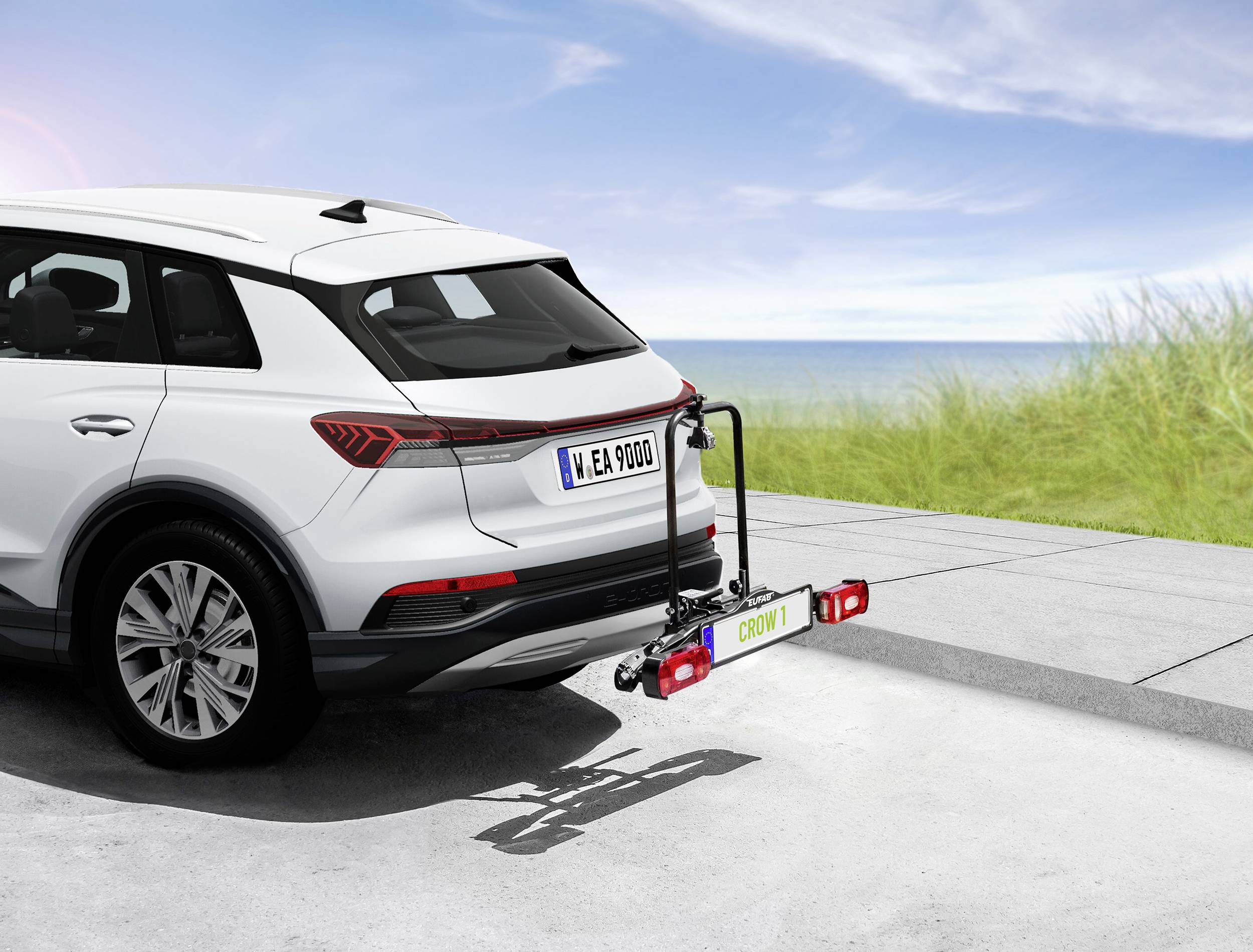 Biały SUV z bagażnikiem rowerowym z tyłu stoi zaparkowany z widokiem na morze i wydmy w tle.