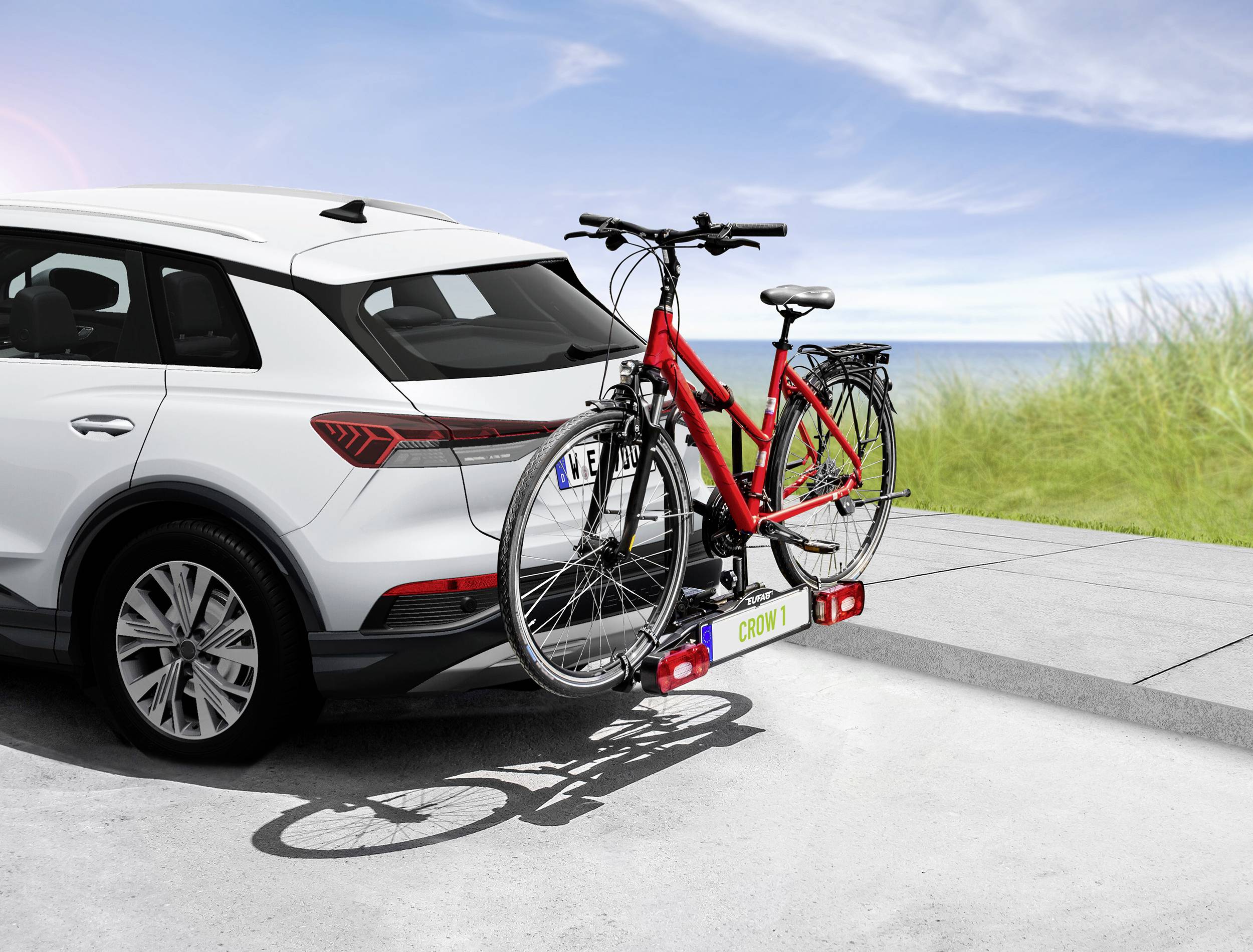 Biały samochód stoi na plaży z czerwonym rowerem przymocowanym do bagażnika na haku. Morze jest widoczne w tle.