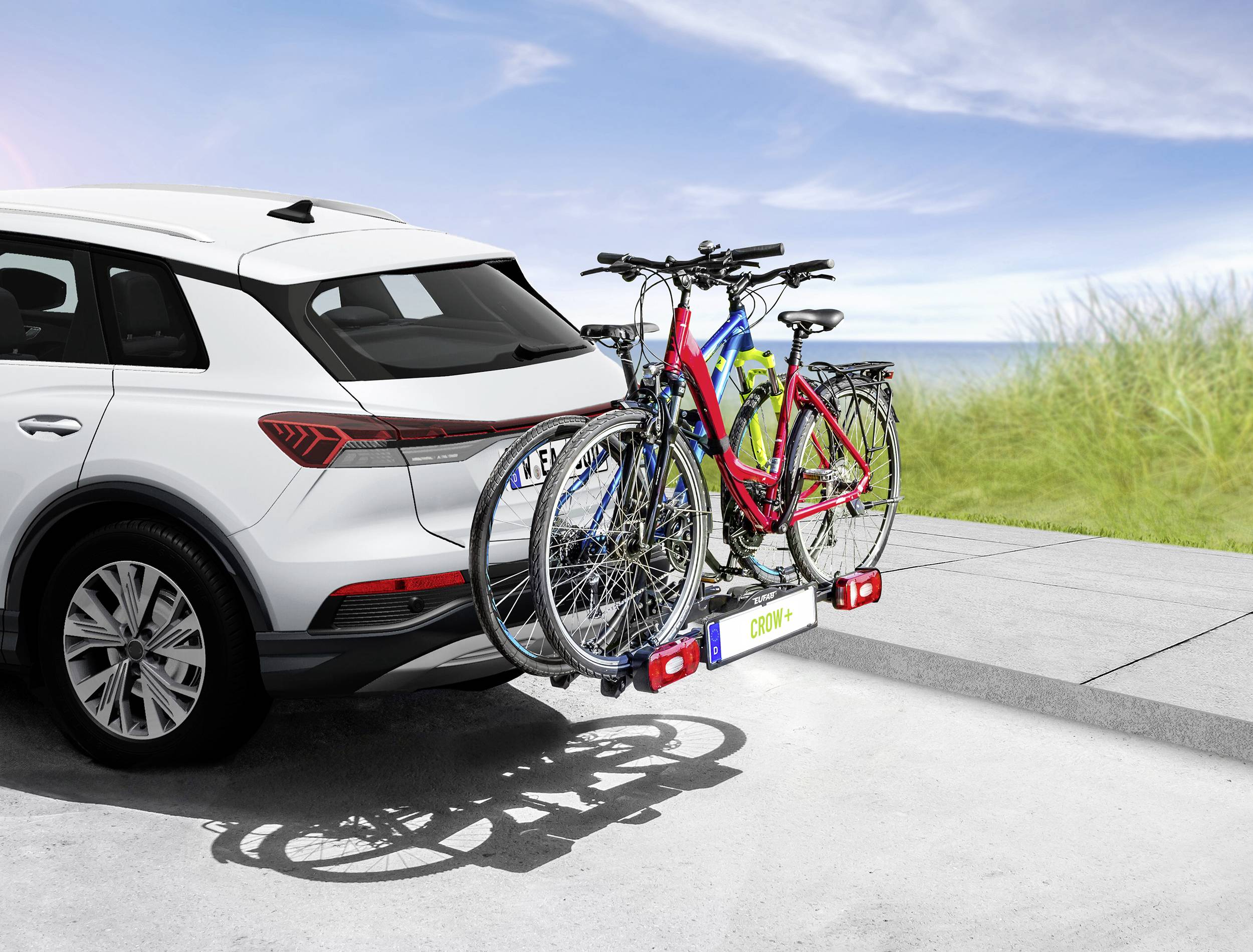 Biały samochód z dwoma rowerami na bagażniku rowerowym z tyłu, zaparkowany na parkingu w słoneczny dzień w pobliżu wybrzeża.