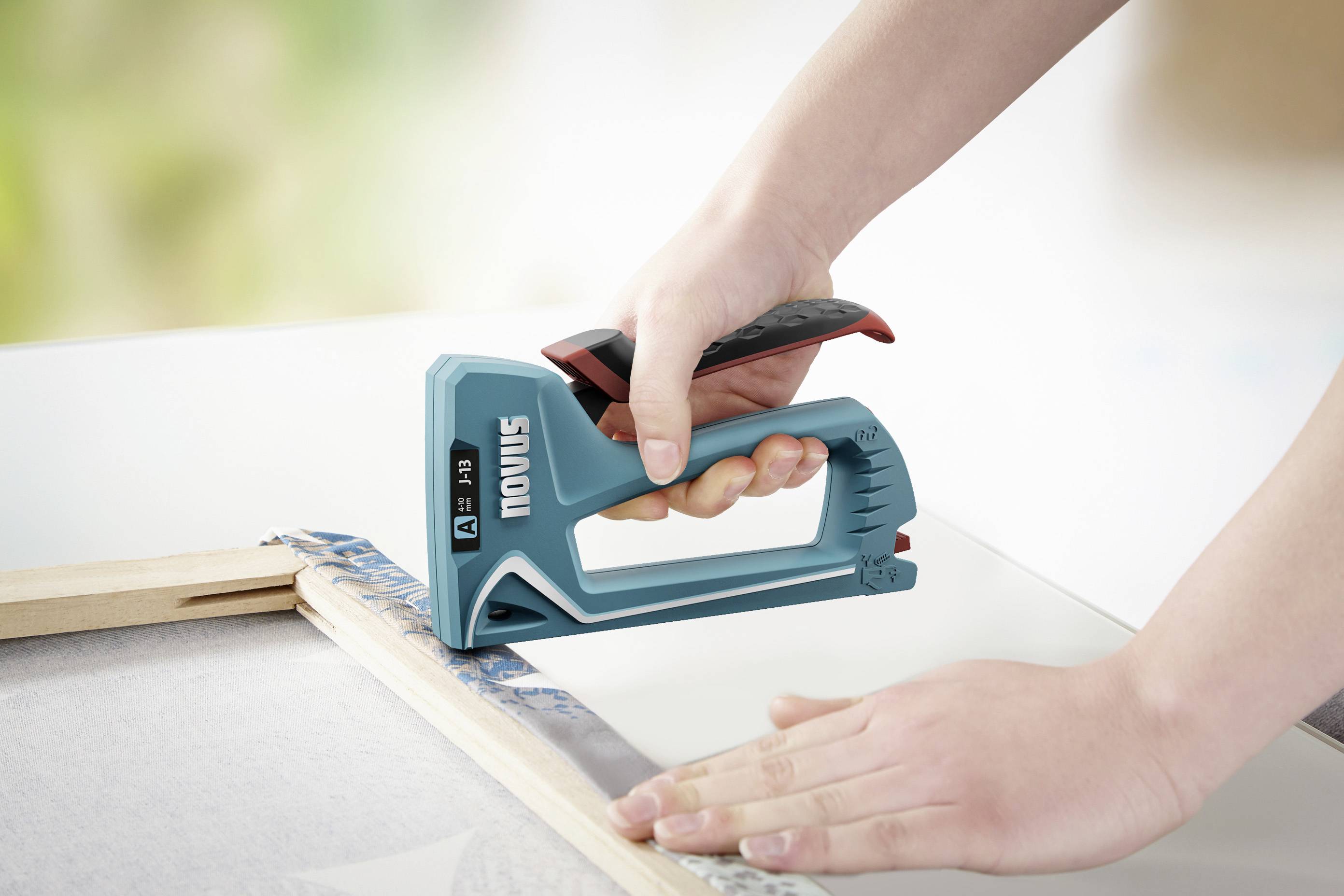 Osoba mocuje folię zszywaczem w kolorze zielonym na drewnianej ramie. Ręce trzymają ramę i kierują zszywaczem.