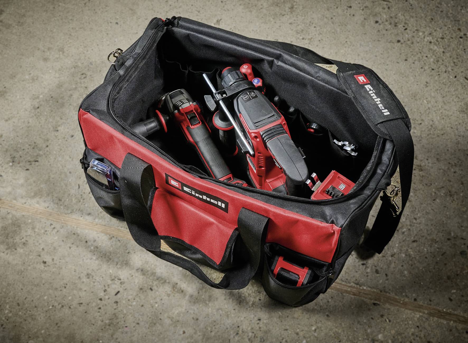 Czerwona torba narzędziowa na betonowej podłodze, wypełniona różnymi narzędziami elektrycznymi.