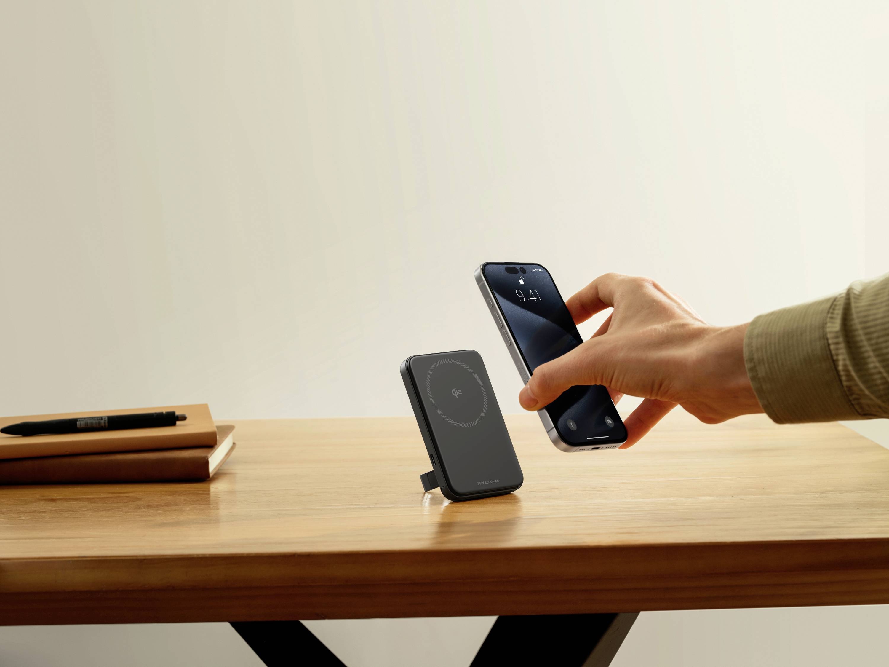 Dłoń trzyma smartfon nad bezprzewodową stacją ładowania na drewnianym stole. W tle notes i długopis.