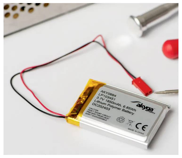 Akumulator litowo-polimerowy z przewodami i czerwonym złączem leży na stole. Tekst na akumulatorze: '1S1P, 1850 mAh, 6,85 Wh, 3,7 V'.