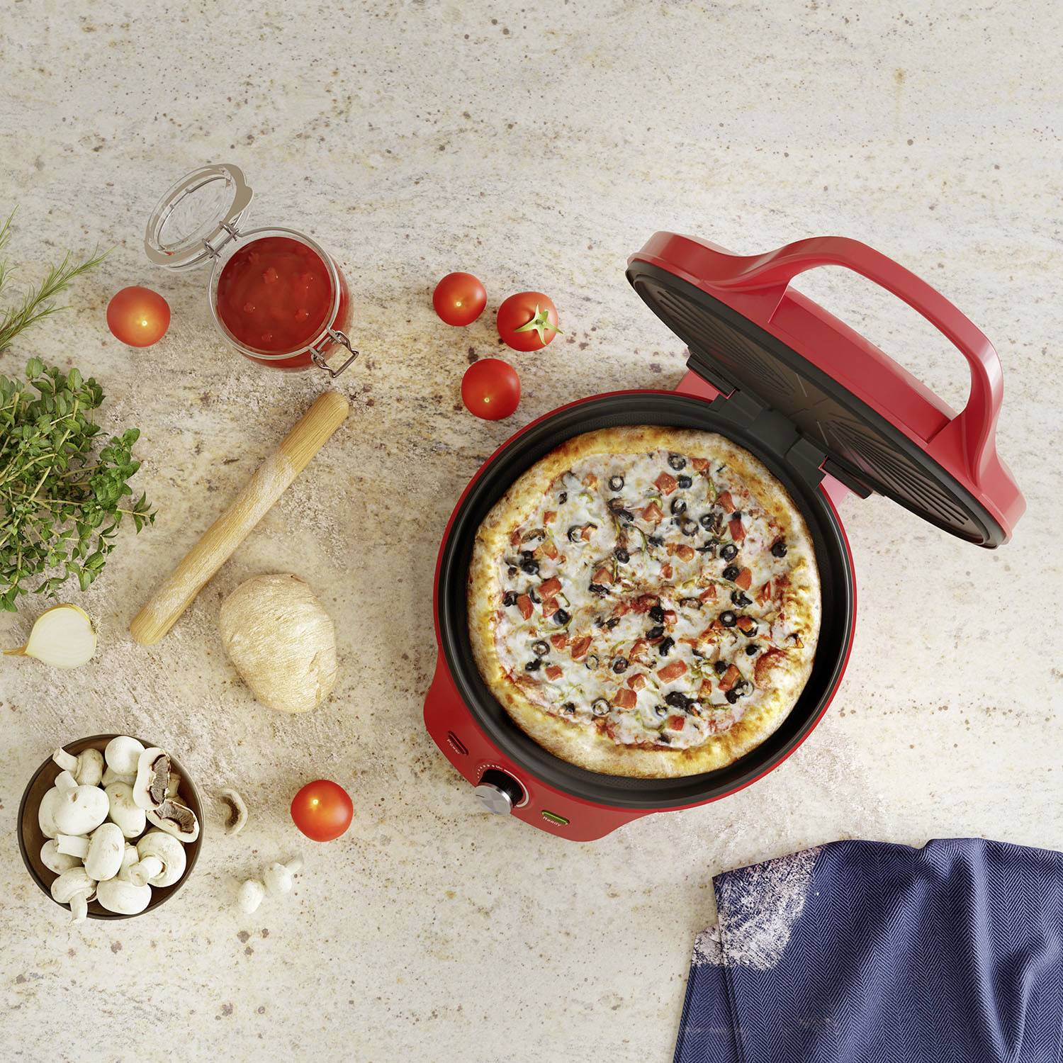 Pizza w czerwonym piecu do pizzy, otoczona pomidorami, pieczarkami, ziołami i słoikiem sosu pomidorowego na stole.