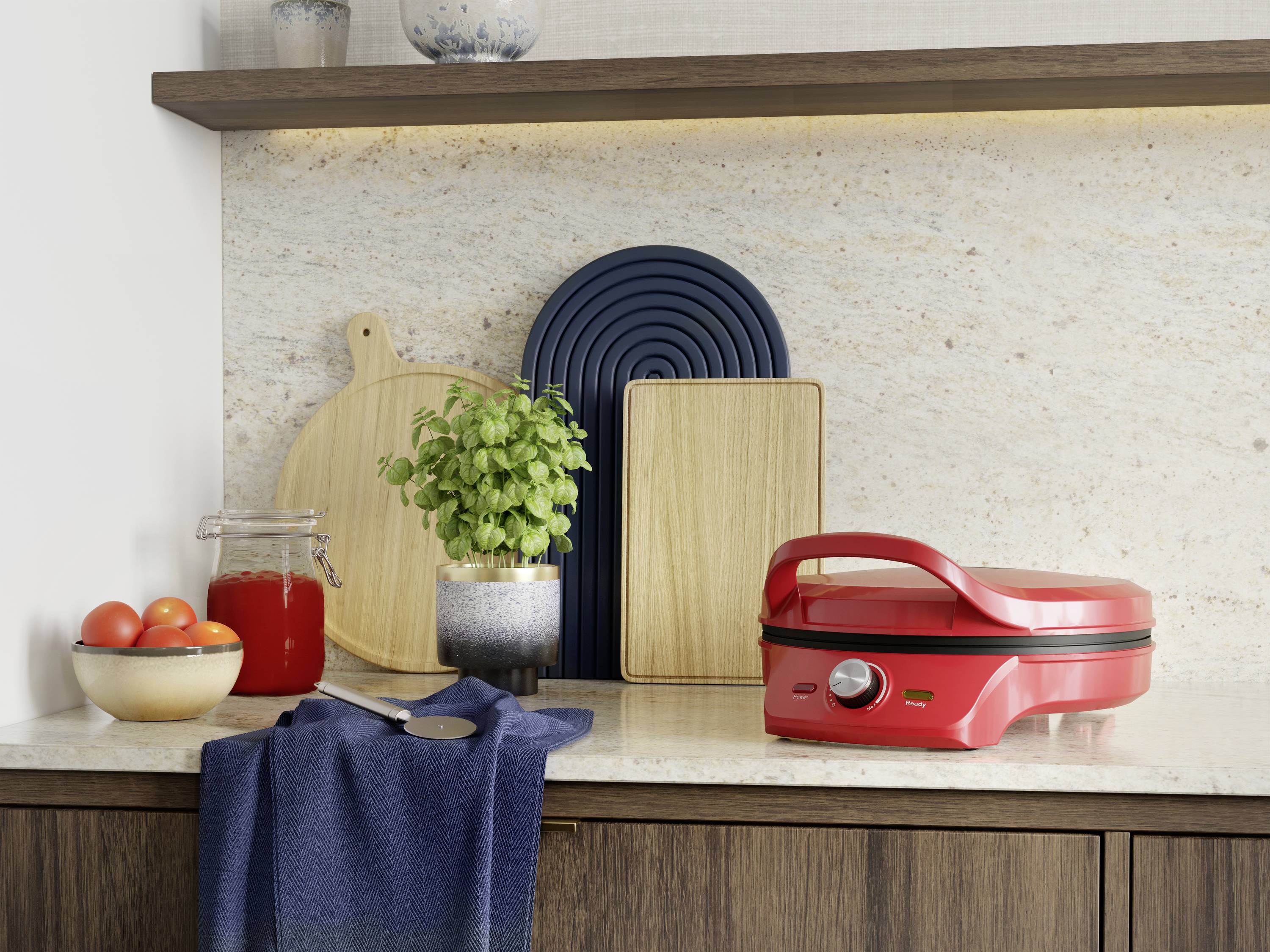 Czerwony grill kontaktowy na blacie kuchennym, otoczony niebieskim płótnem, świeżymi pomidorami i doniczką bazylii na tle desek do krojenia.