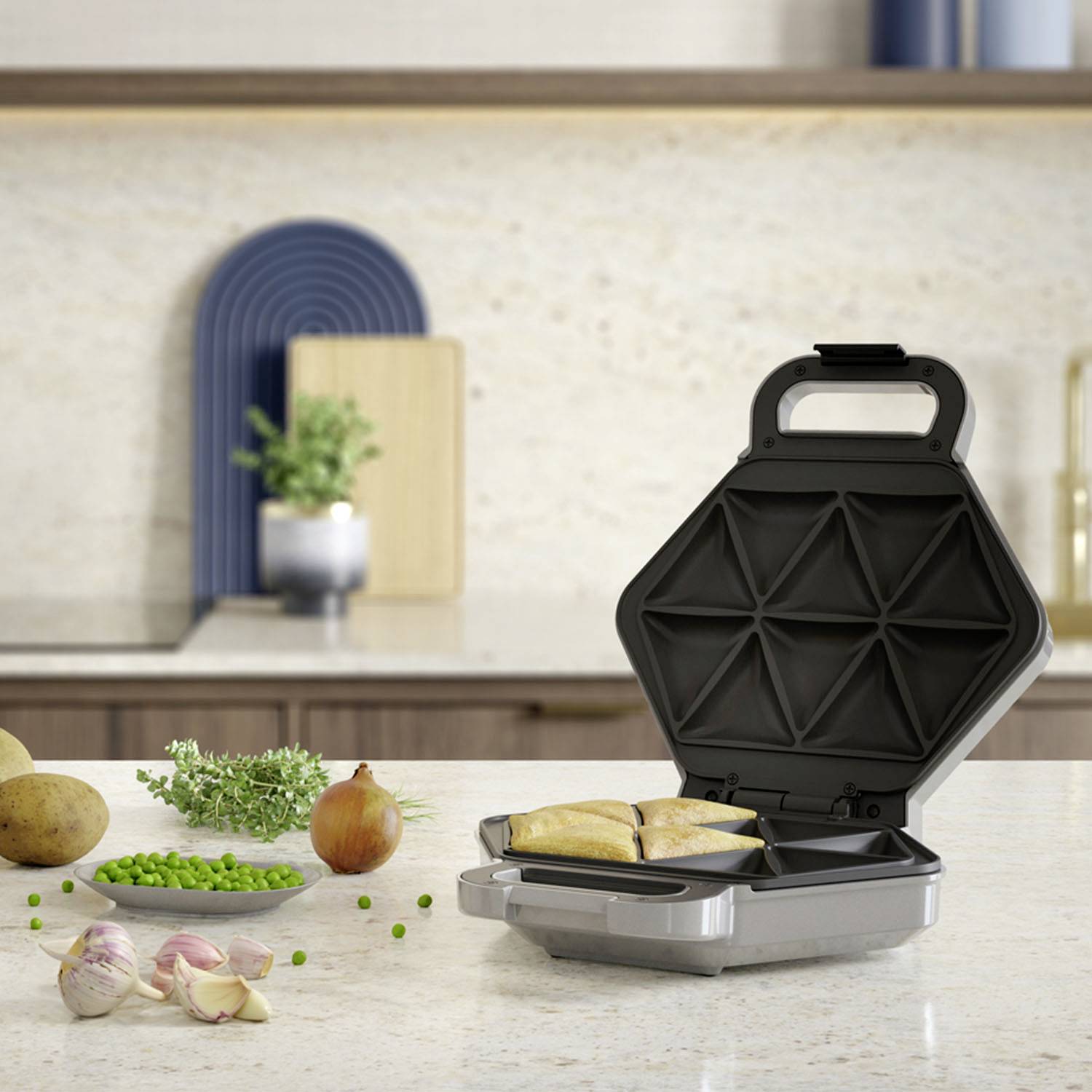 Otwarty toster do kanapek stoi na blacie kuchennym, a w tle widoczne są składniki takie jak cebula, ziemniaki i groszek.