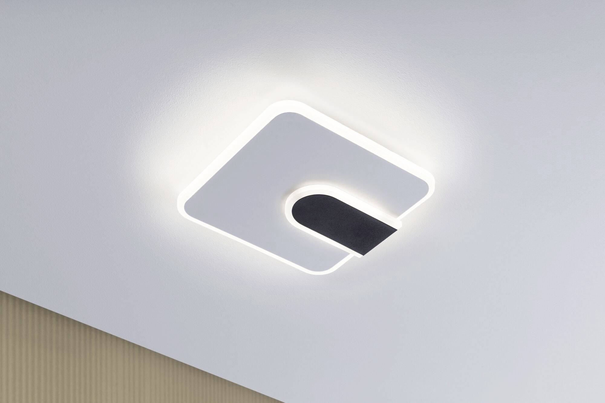 Nowoczesna lampa sufitowa o kwadratowym kształcie z zakrzywionym czarnym elementem pośrodku, która tworzy miękkie, rozproszone światło.