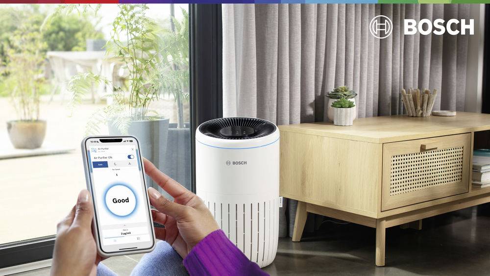 Osoba sprawdza jakość powietrza na smartfonie. Oczyszczacz powietrza marki Bosch stoi obok w nowoczesnym salonie.