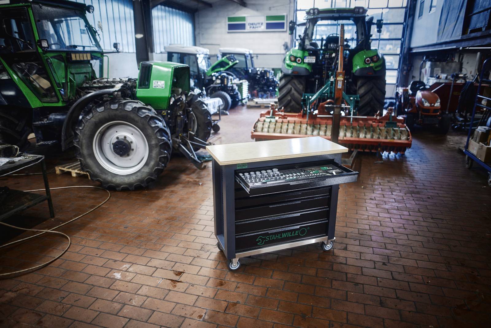 Warsztat z kilkoma traktorami i wózkiem narzędziowym pośrodku, otoczony jasnym światłem i różnymi maszynami.