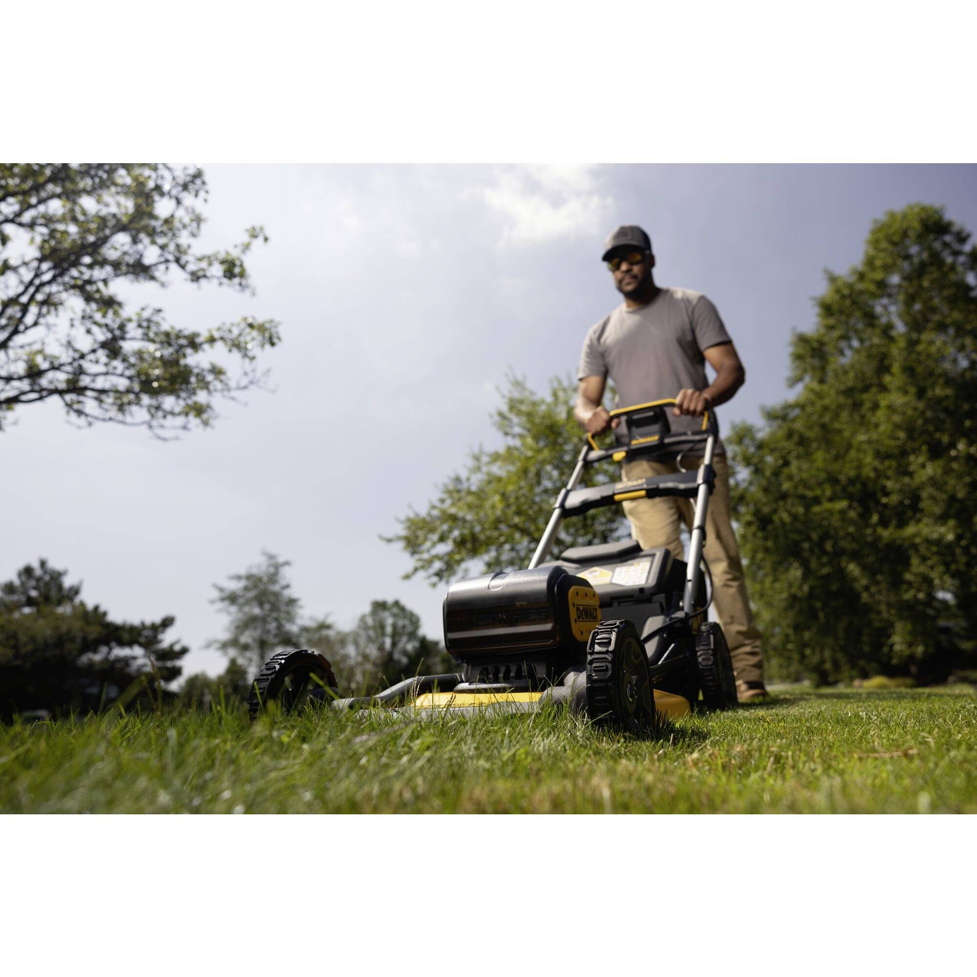 Mężczyzna kosi trawę żółto-czarną kosiarką w słoneczną pogodę. W tle rosną drzewa, a niebo jest częściowo zachmurzone.