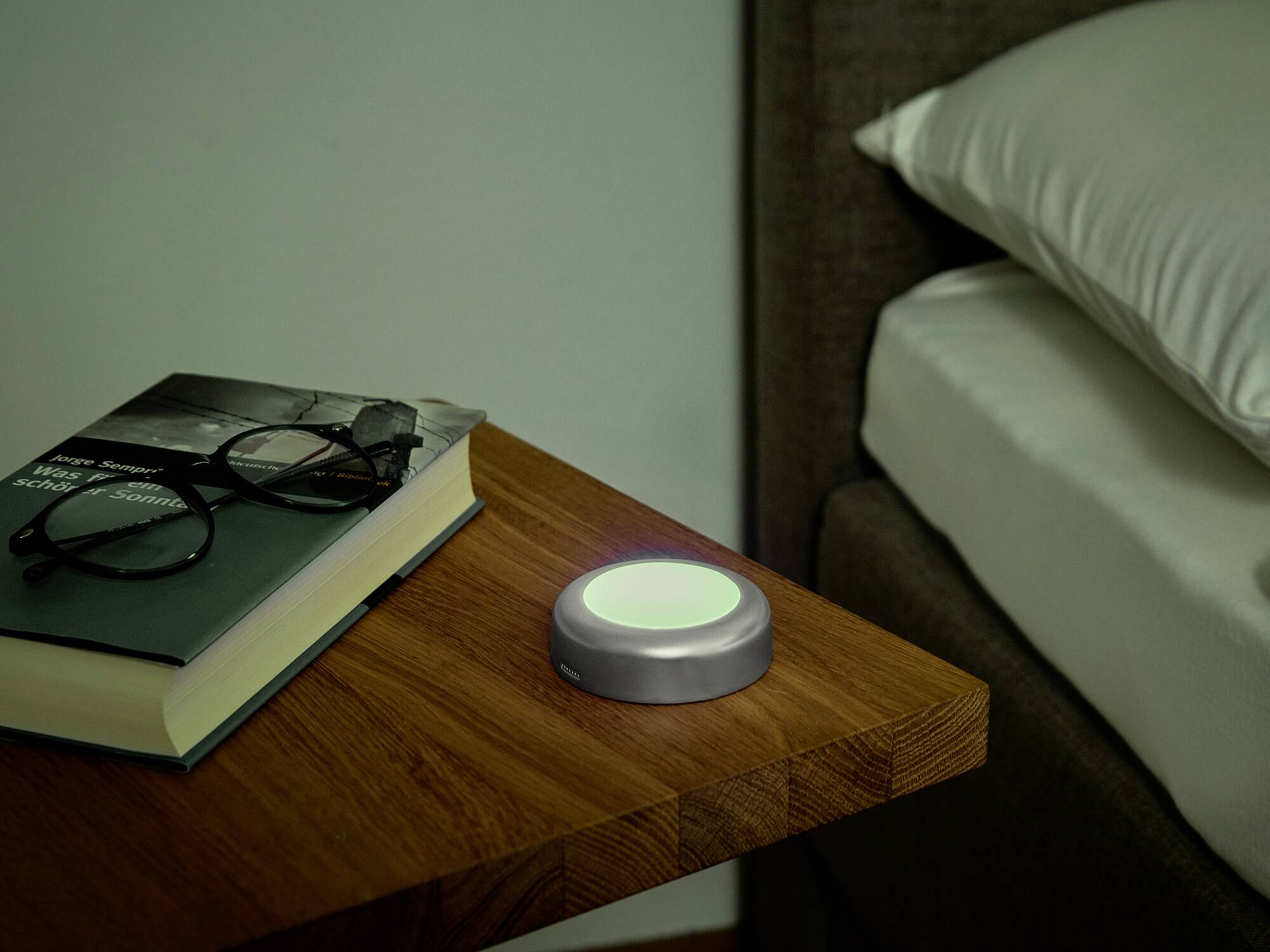 Na stoliku nocnym leży książka i okulary obok świecącej, okrągłej lampki nocnej. Obok stoi łóżko.