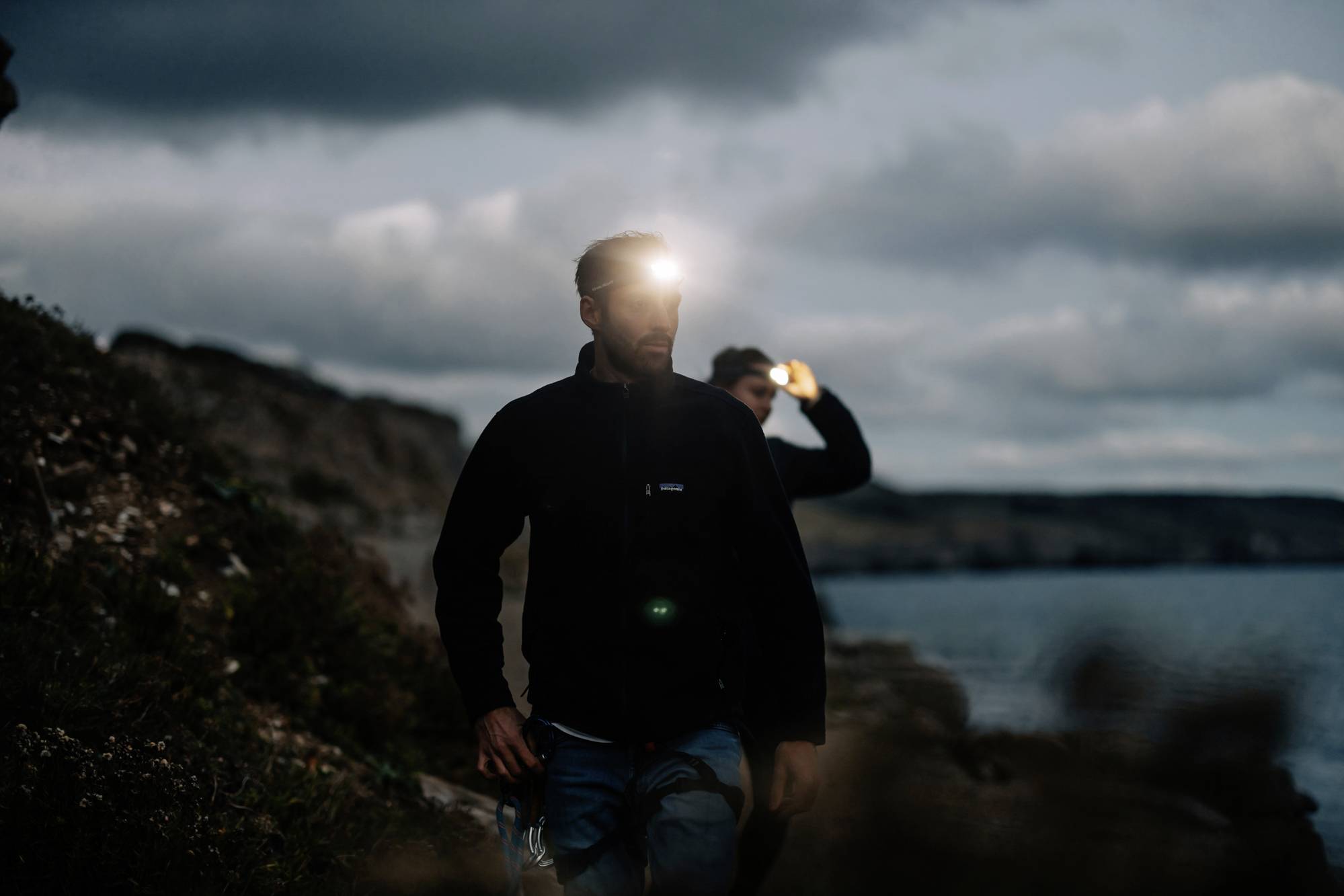 Dwie osoby idą wzdłuż wybrzeża w ciemności. Noszą czołówki, które oświetlają ich otoczenie. Niebo jest zachmurzone.