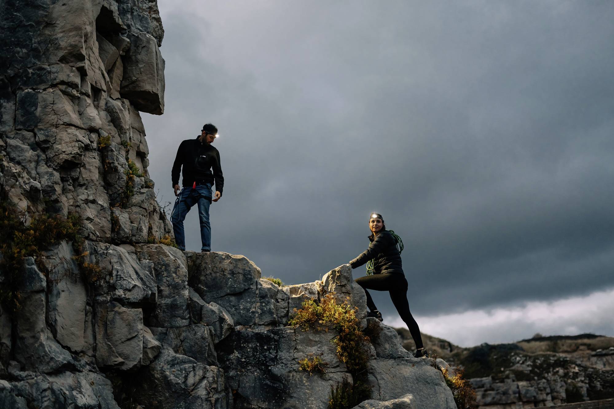 Dwie osoby wspinają się na skalistą skałę pod pochmurnym niebem. Scena oddaje ducha przygody i fizyczne wyzwanie w środowisku naturalnym.