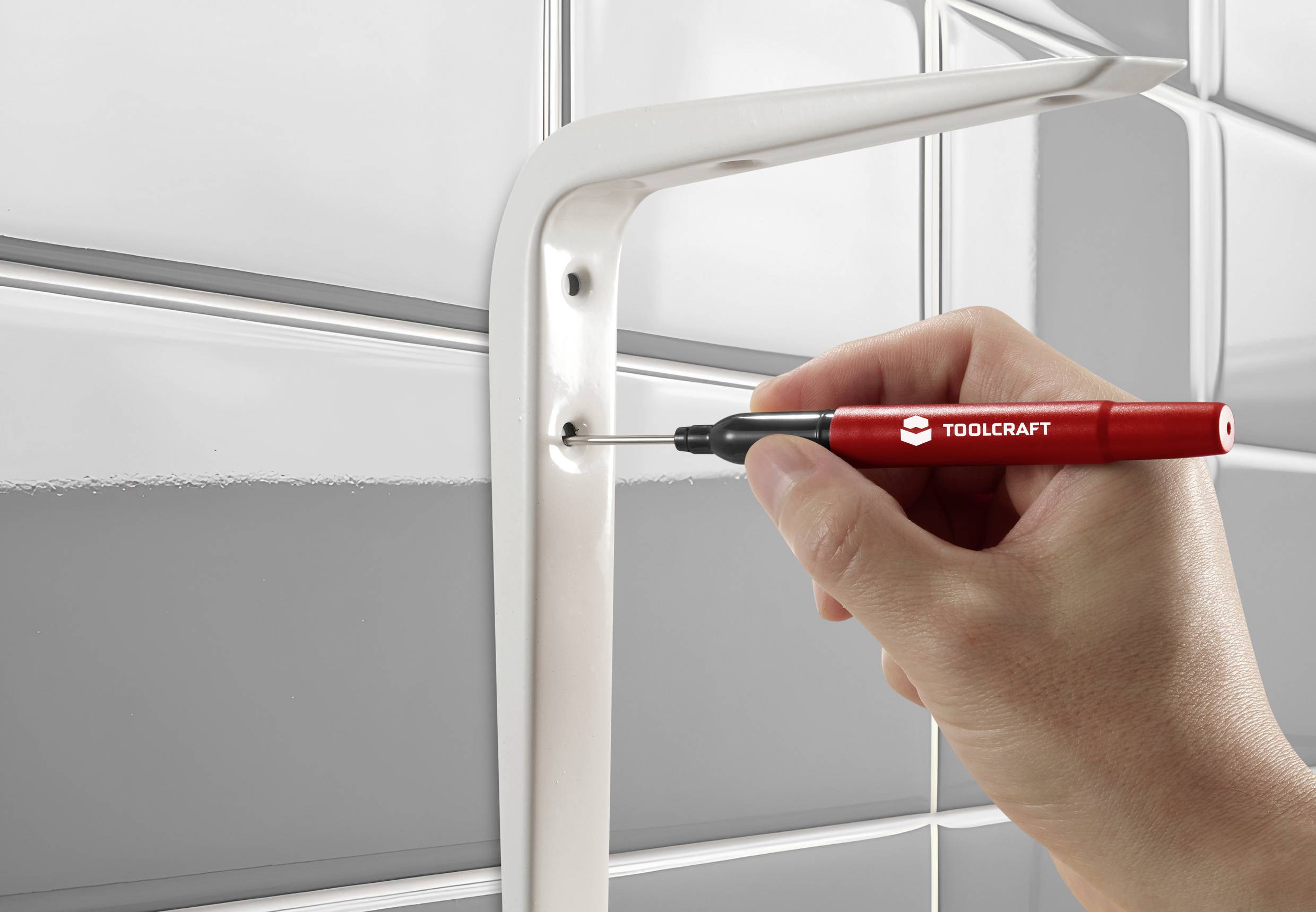 Jedna ręka trzyma czerwony wkrętak TOOLCRAFT i dokręca śrubę w białym uchwycie, aby zamocować go na ścianie.