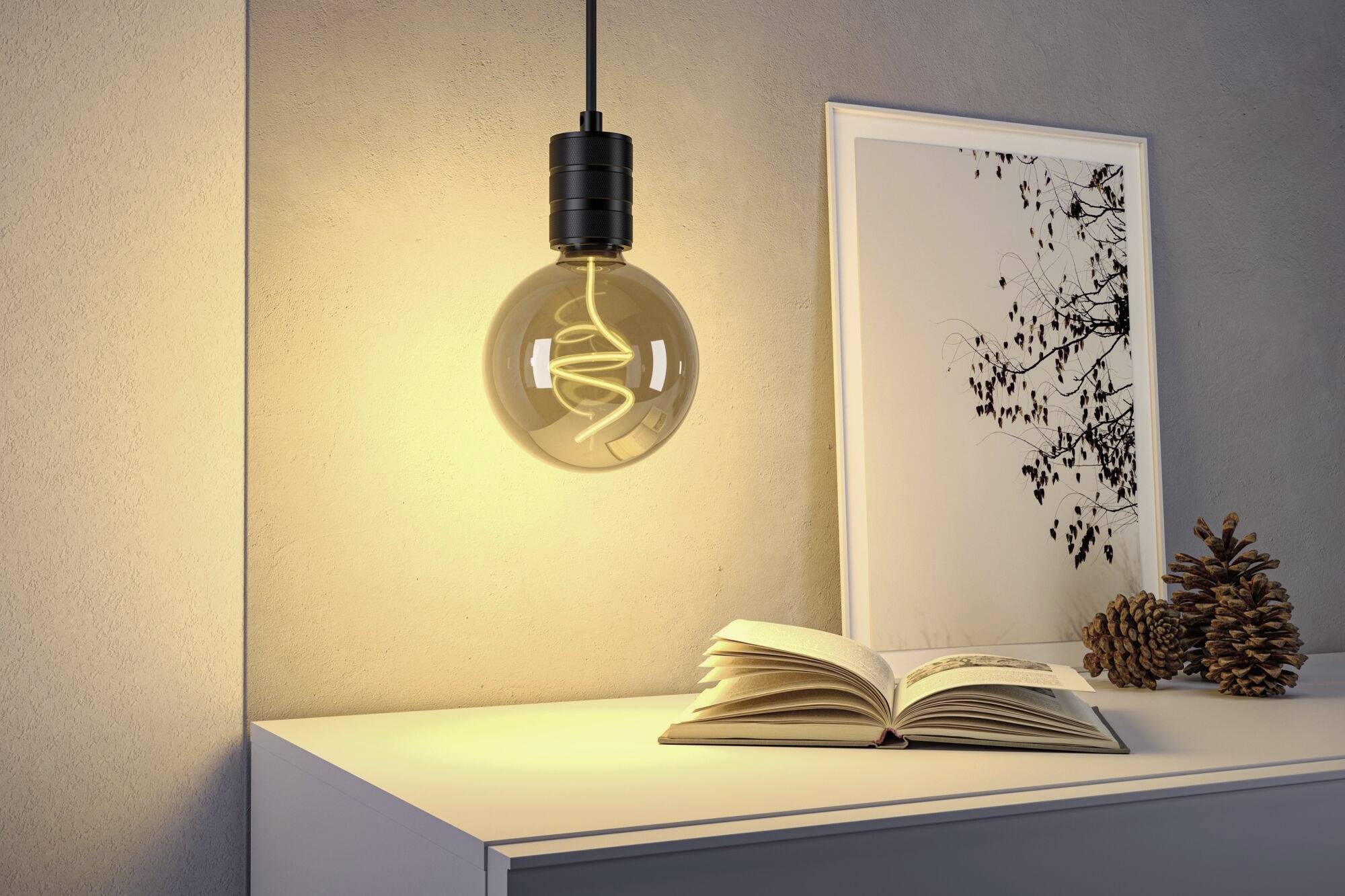 Świecąca żarówka wisi nad białym stołem, na którym leży otwarta książka, oprawione zdjęcie i szyszki.