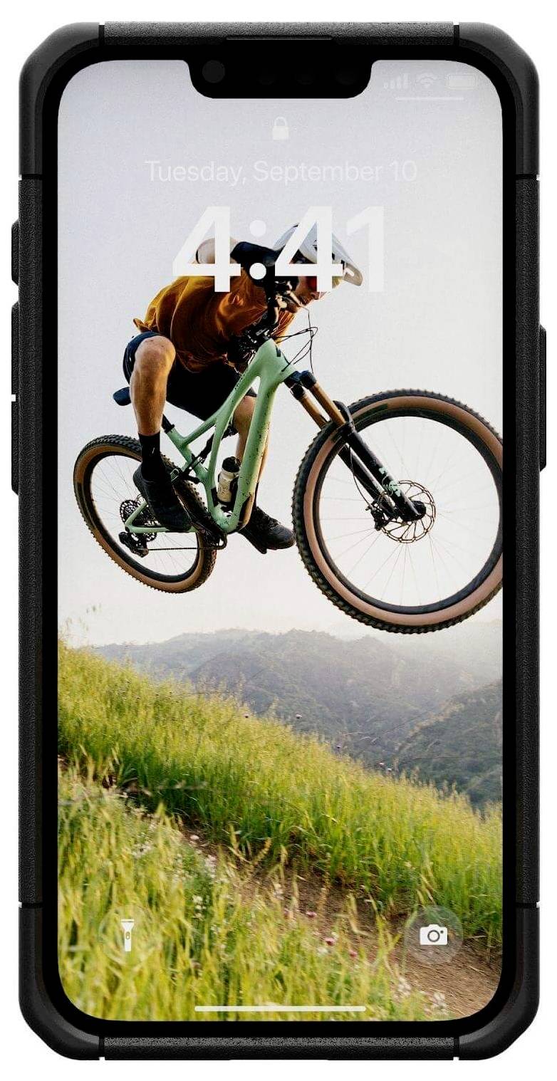 Rowerzysta górski skacze na rowerze przez wzniesienie. W tle widać zielone łąki i góry.