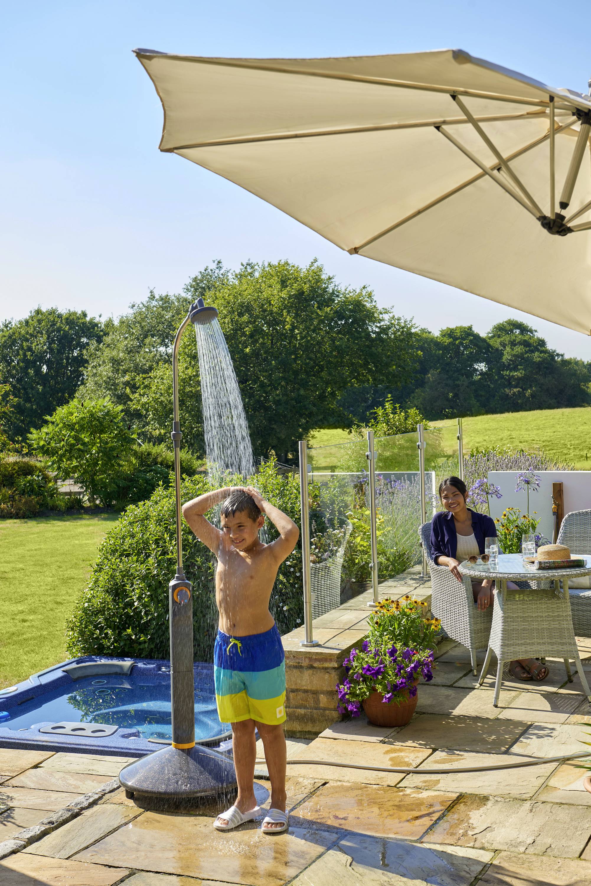 Dziecko bierze prysznic na tarasie pod gołym niebem, podczas gdy siedząca osoba przypatruje się spod parasola. W tle widoczny jest ogród.