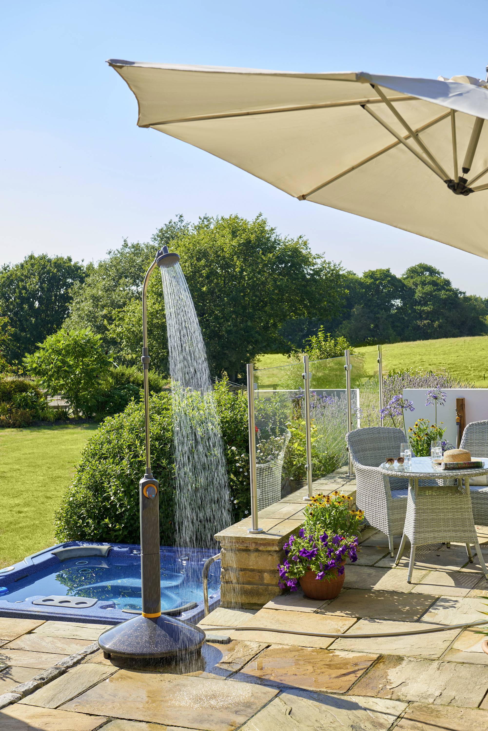 'Ogród z leżakiem, parasolem, zestawem mebli ogrodowych i prysznicem ogrodowym. W tle jacuzzi i zielony krajobraz.'