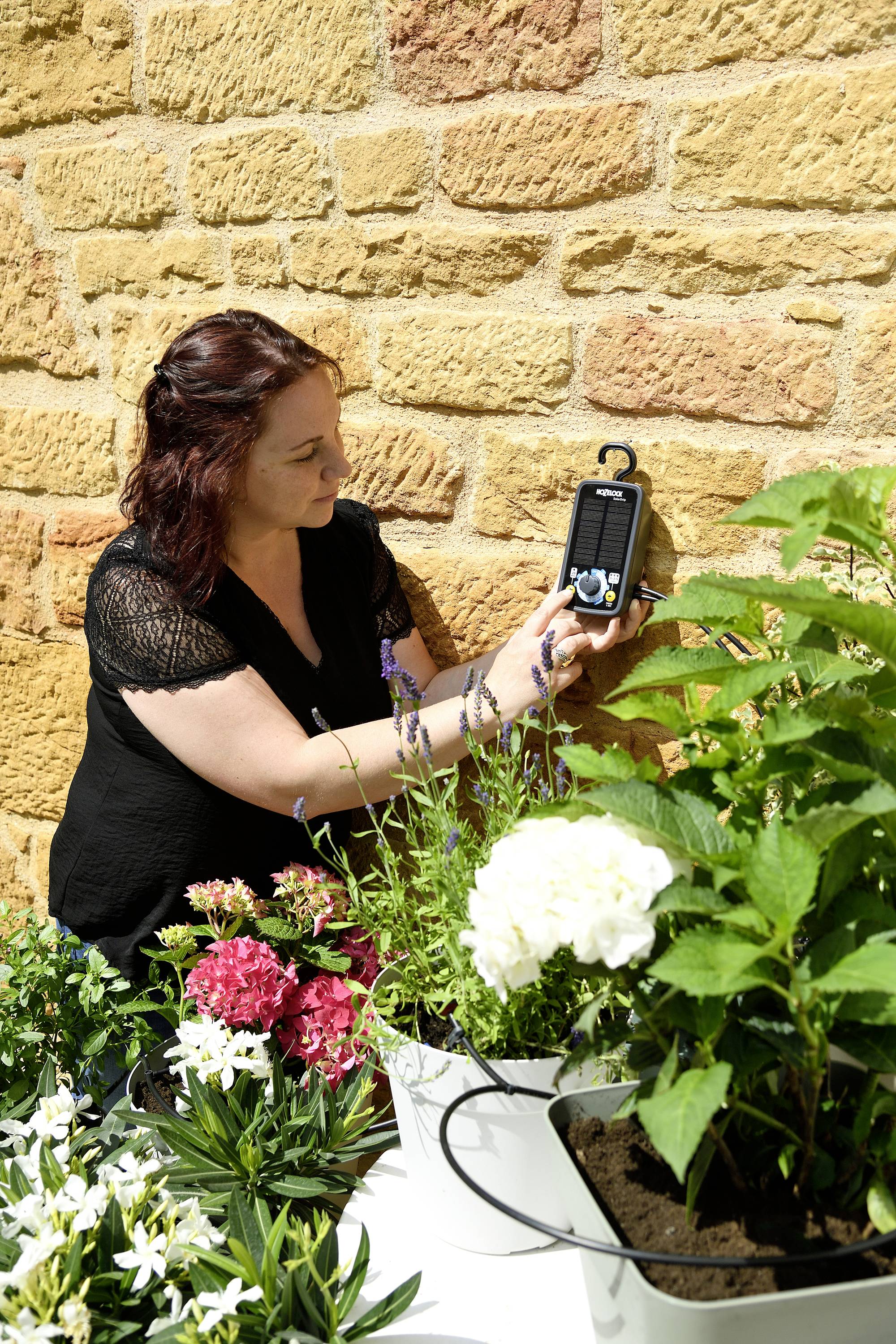 Kobieta sprawdza urządzenie przy kamiennej ścianie, otoczona kwitnącymi roślinami w doniczkach. Słoneczna pogoda.