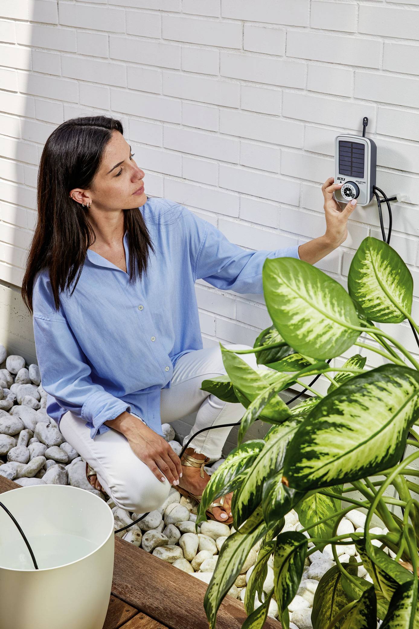 Kobieta w niebieskiej bluzce sprawdza urządzenie zamontowane na ścianie obok rośliny. Znajduje się w jasnym pomieszczeniu z kamykami.