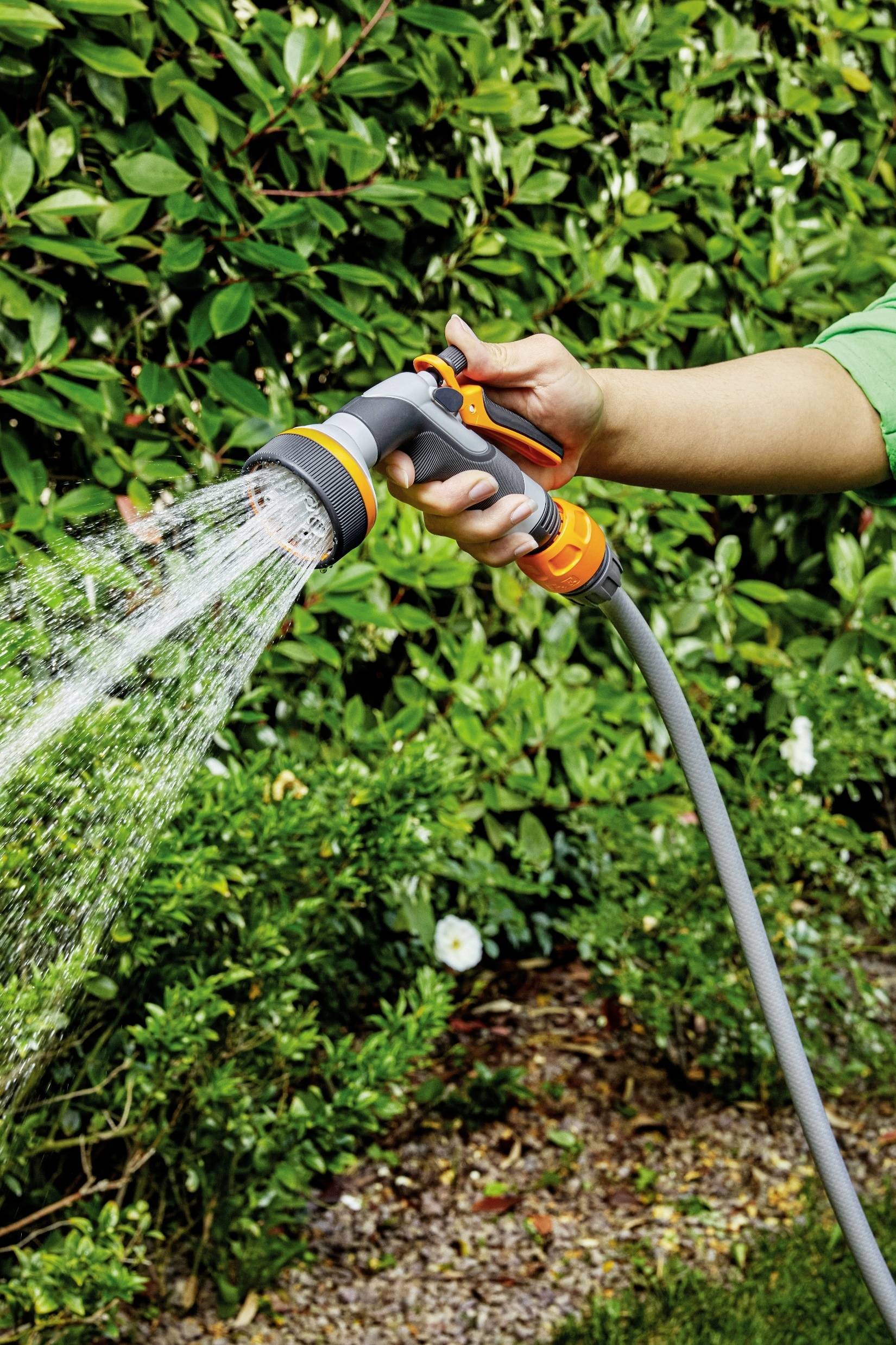Osoba podlewa rośliny wężem ogrodowym w ogrodzie. W tle widoczne są zielone krzewy.
