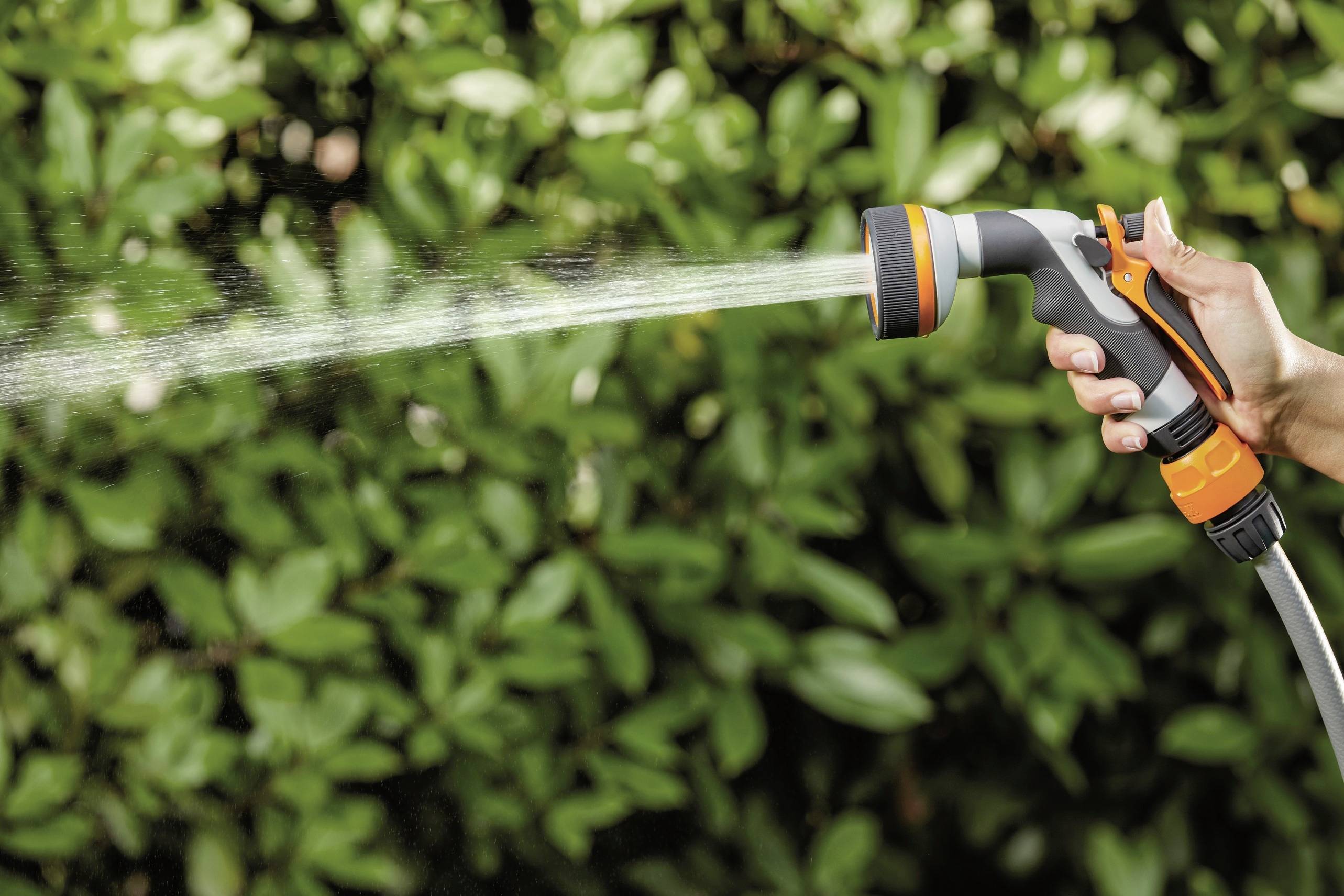 Ręka trzyma wąż ogrodowy z dyszą natryskową, nawadniając zielone liście w tle.