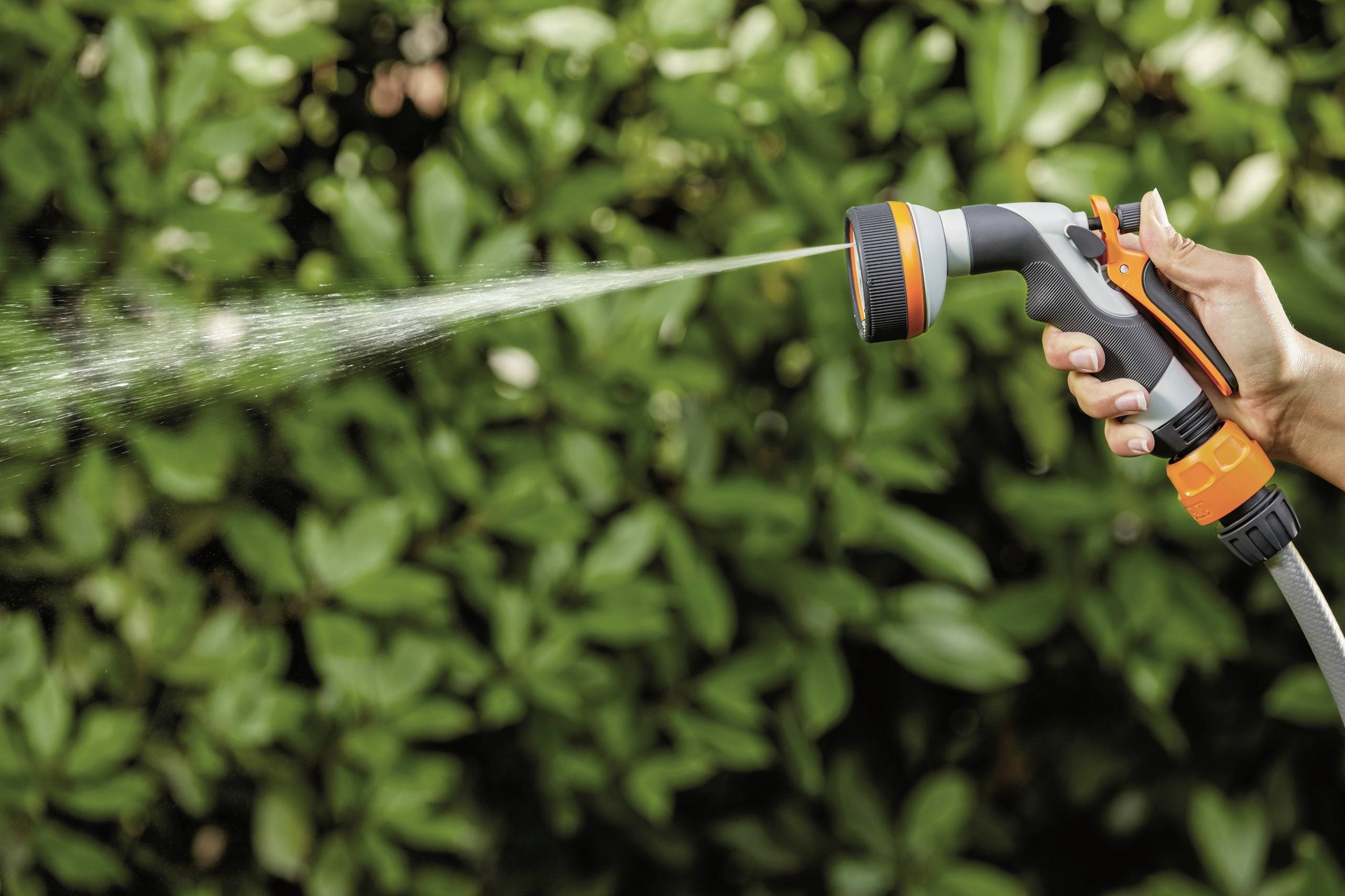 Ręka trzyma wąż ogrodowy z dyszą, z której tryska woda, na tle zielonych liści.