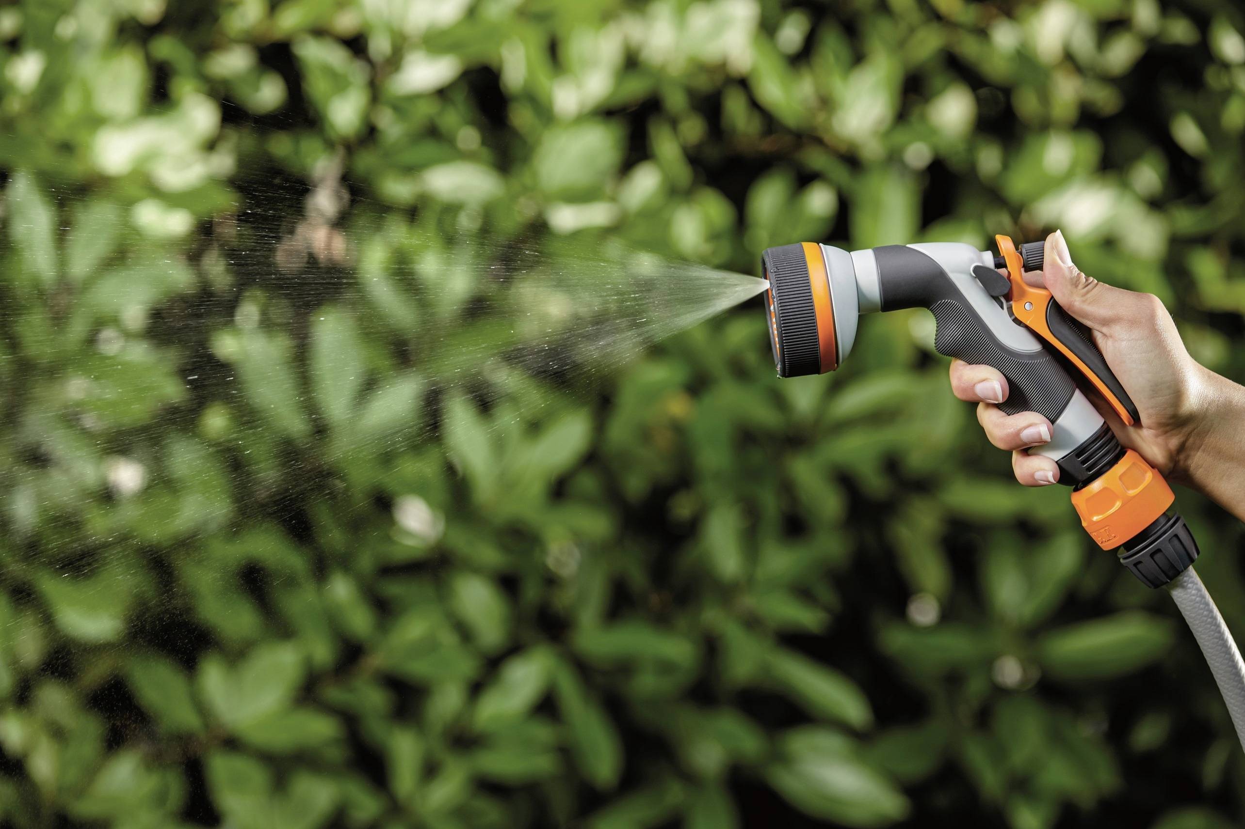 Dłoń trzyma dyszę ogrodową, która rozpyla wodę na zielone liście. Tło rozmyte, pełne gęstej, zielonej roślinności.