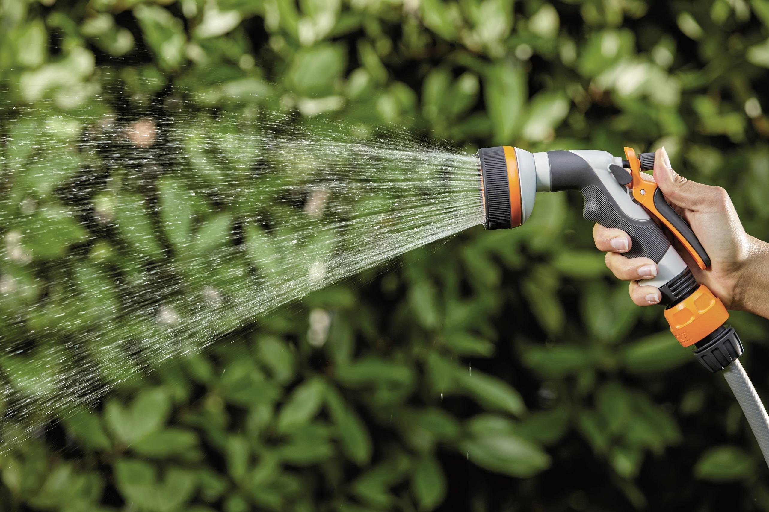 Ręka trzyma pomarańczowo-szarą dyszę rozpylającą i nawadnia rośliny na rozmytym zielonym tle.