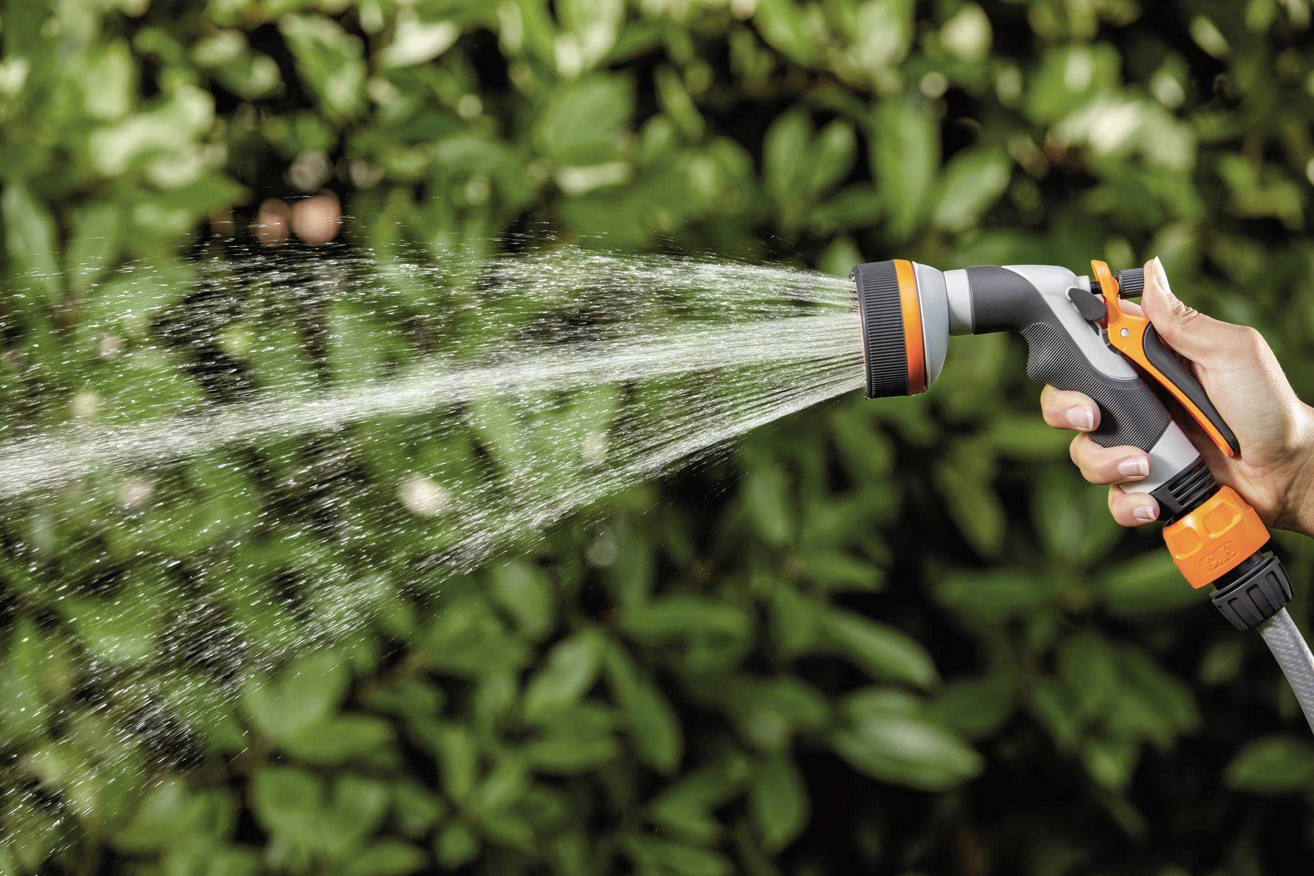 Dłoń trzyma wąż ogrodowy i rozpyla wodę na zielone liście w tle.