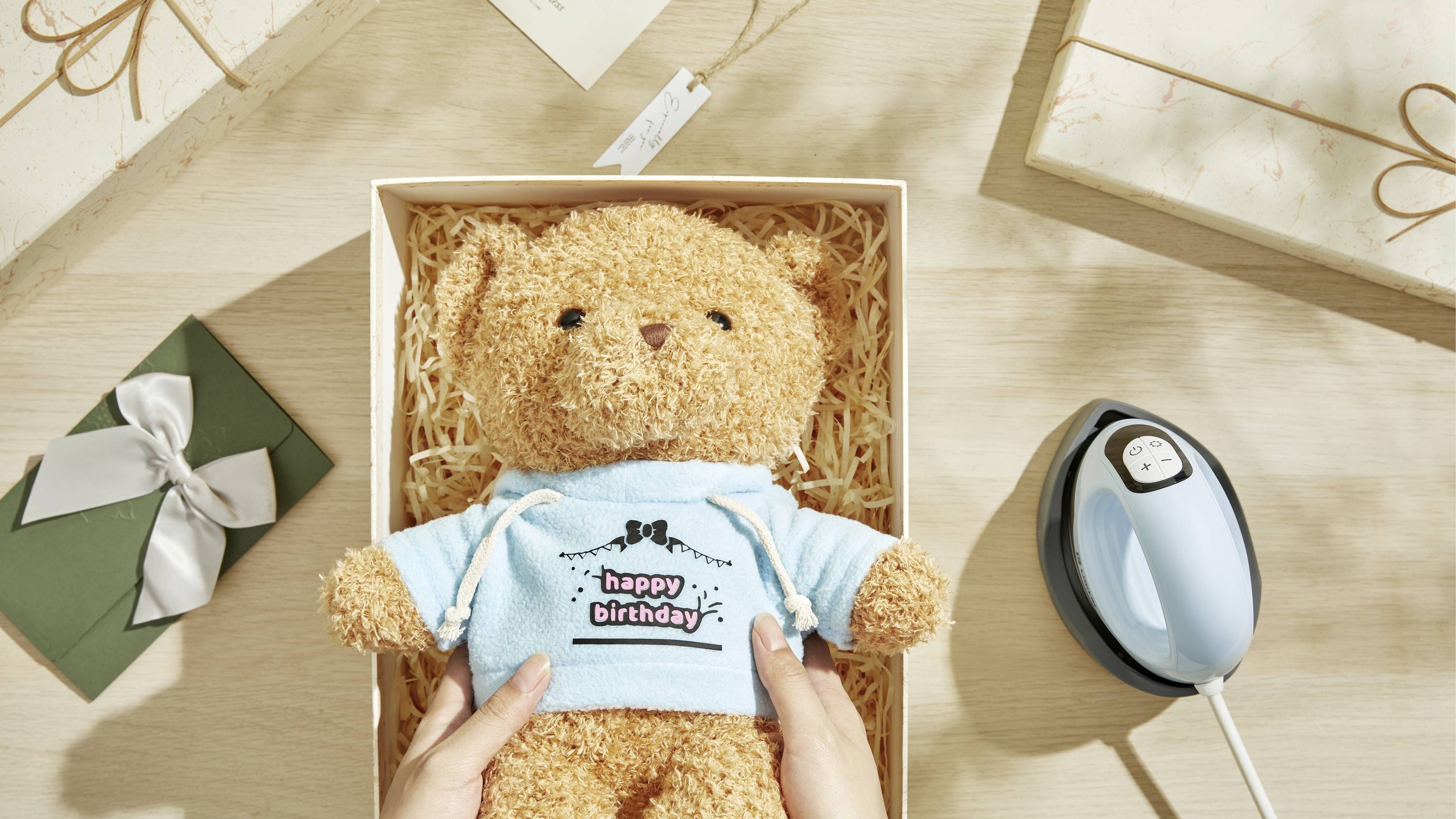 Pluszowy miś w otwartym pudełku prezentowym, ubrany w niebieski sweter z napisem 'happy birthday'; otoczony prezentami i żelazkiem.